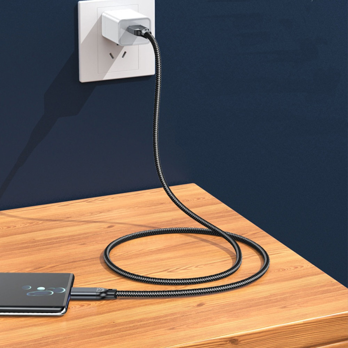 Ein schwarzes Ladekabel verbindet ein Netzteil mit einem Smartphone auf einem Holzschreibtisch.