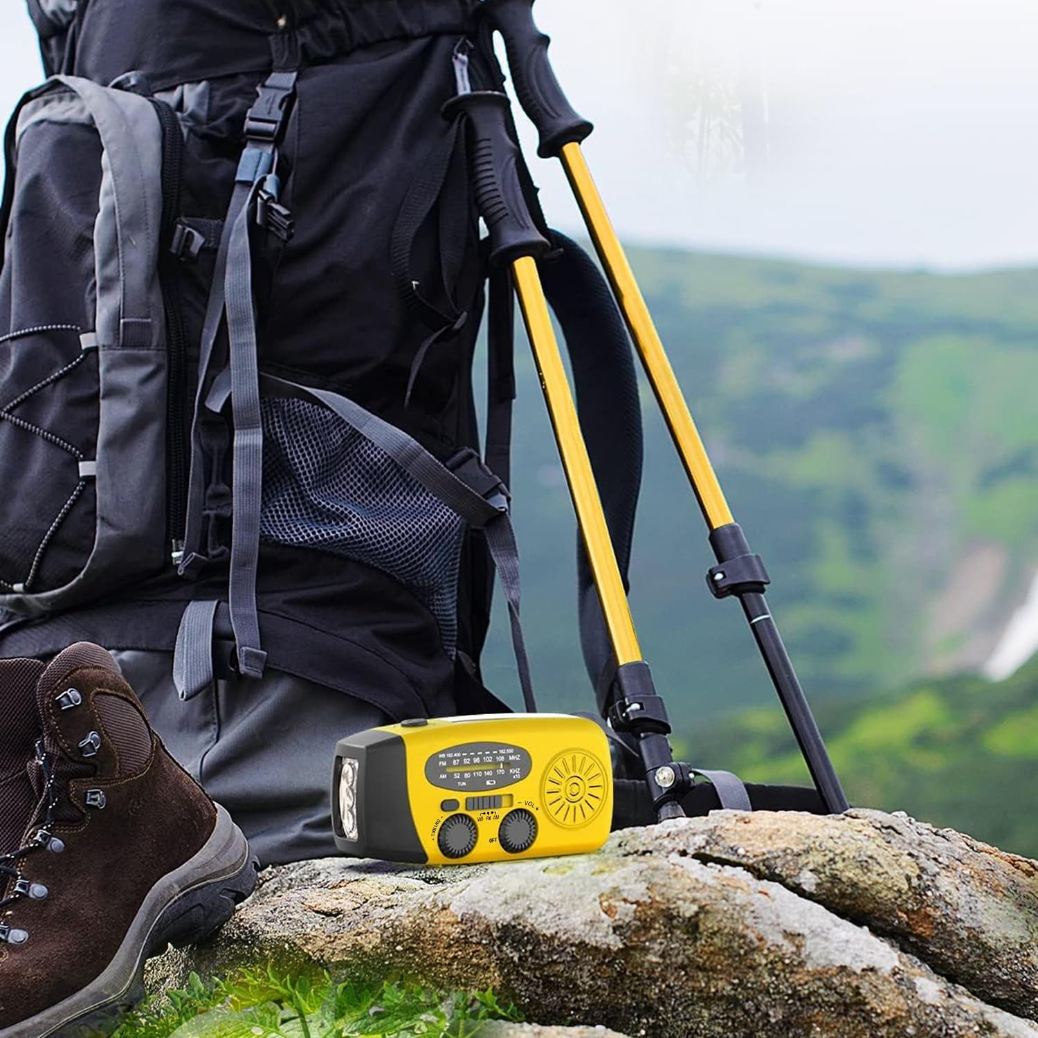 Ein gelbes Notfallradio auf einem Stein mit Rucksack und Wanderstöcken im Hintergrund in einer Berglandschaft.