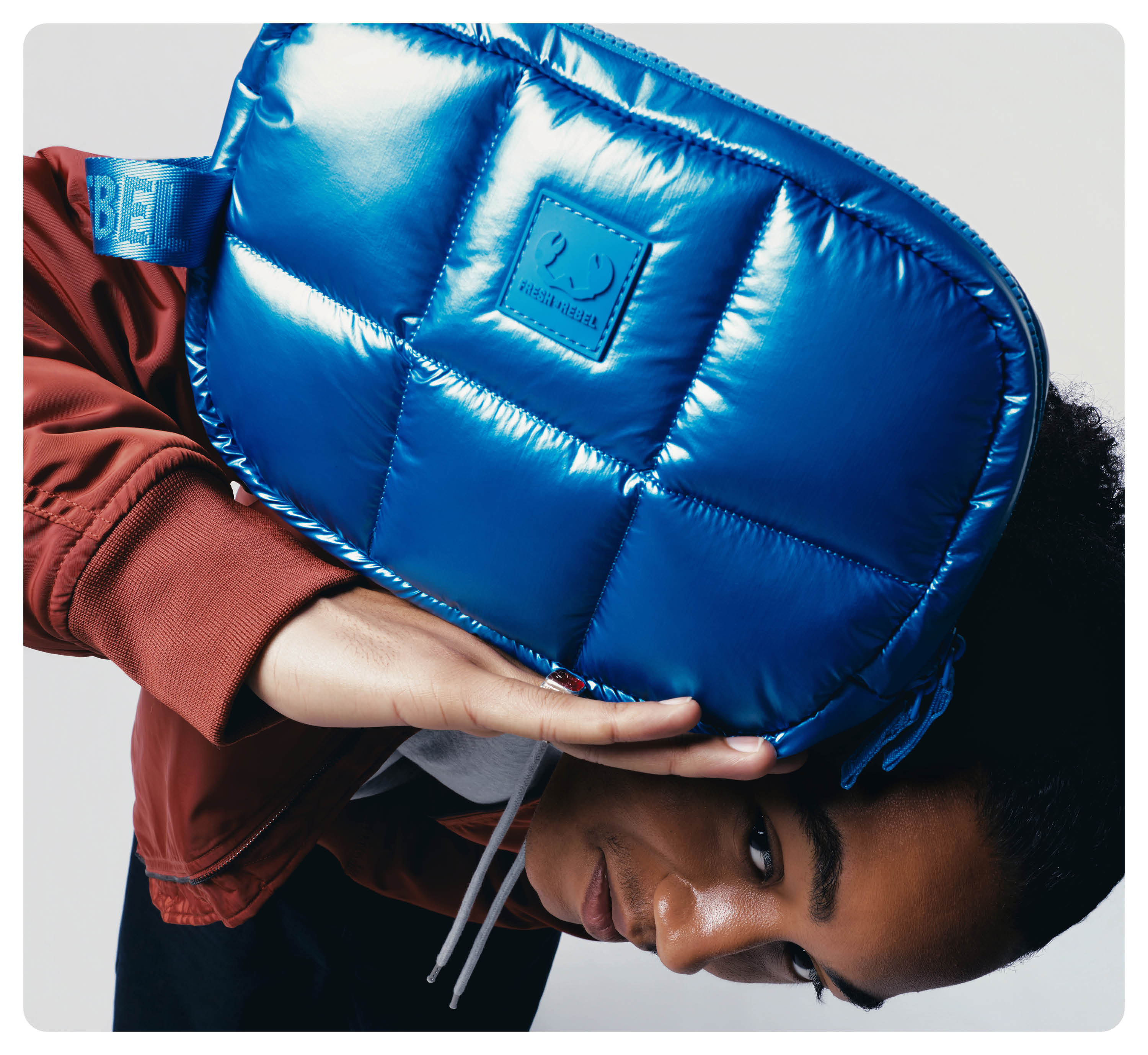 Eine Person in einer rostfarbenen Jacke hält vor hellem Hintergrund eine glänzende blaue Stepptasche an den Kopf.