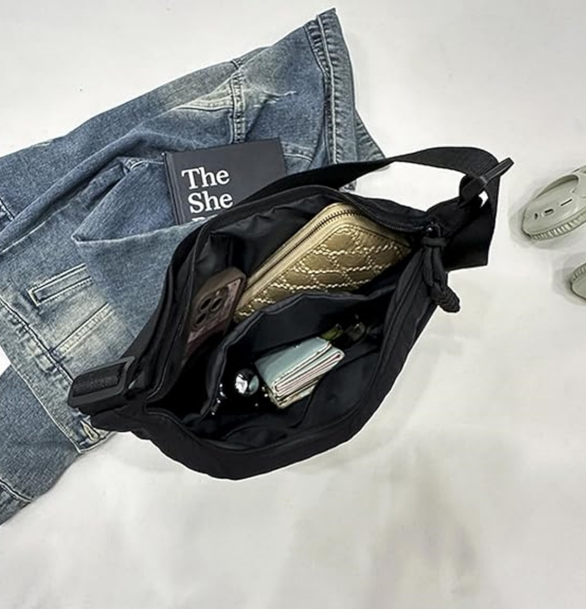 Eine offene schwarze Umhängetasche mit goldener Geldbörse, Handy und Buch auf weißer Oberfläche neben Jeansjacke.