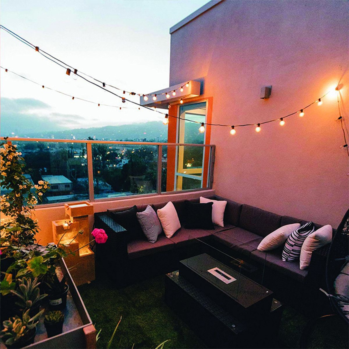 Gemütliche Dachterrasse mit Lichterketten, grauem Sofa, Kissen und Pflanzen bei Dämmerung.