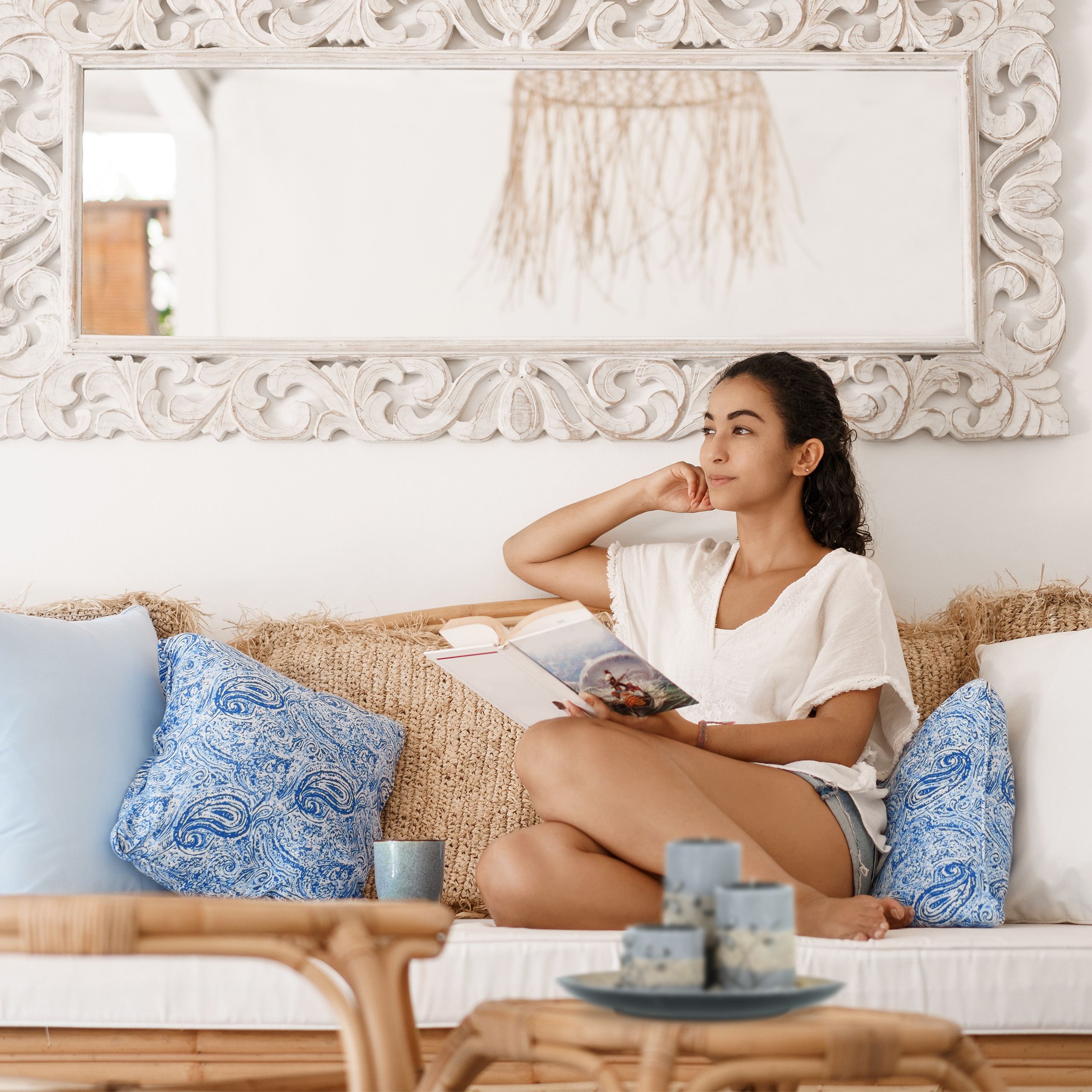 Eine Frau in weißer Bluse sitzt auf einem Sofa und liest ein Buch vor einem verzierten Spiegel.