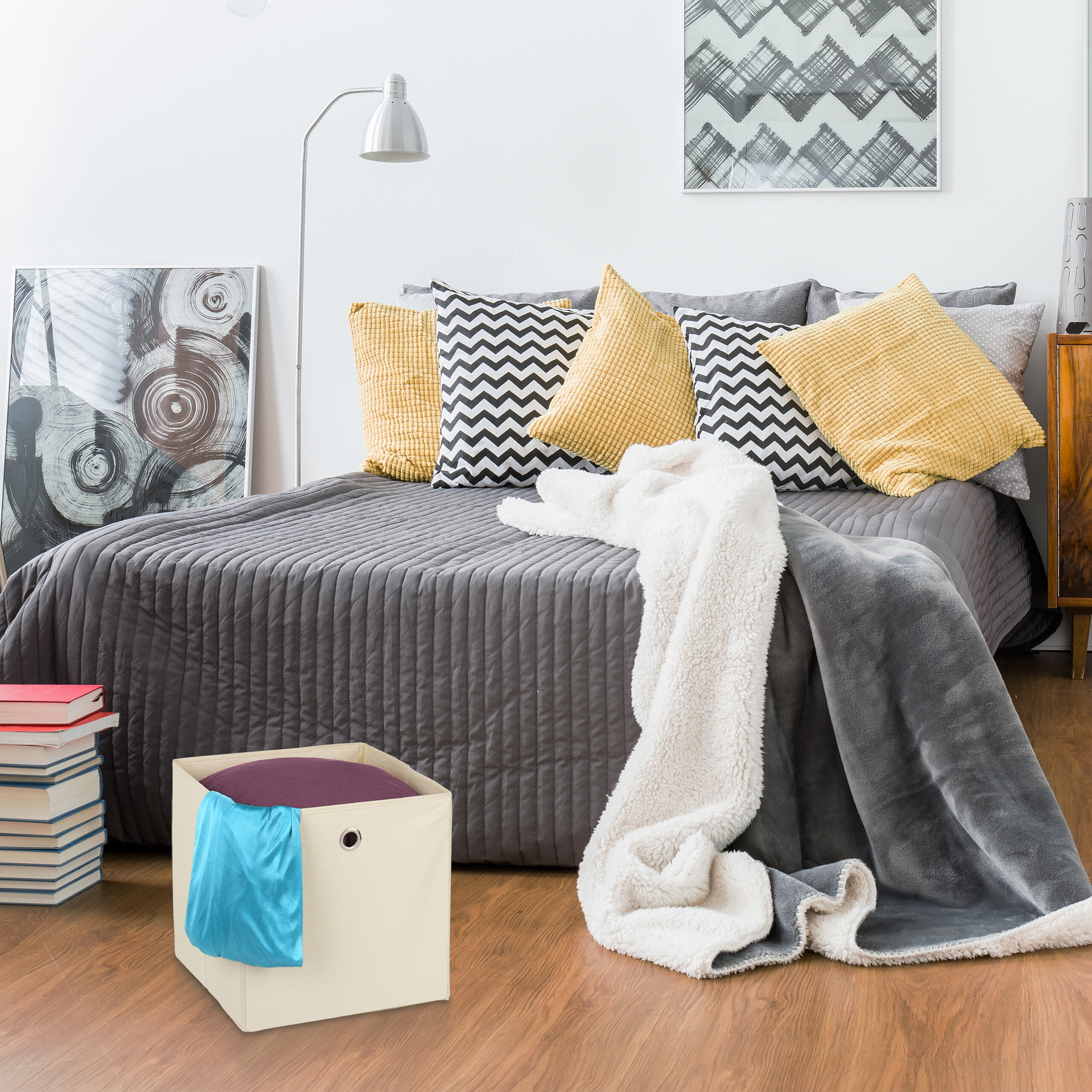 Gemütliches Schlafzimmer mit grauem Bett, gelben Kissen, Decken, Lampe, Bildern und einer Aufbewahrungsbox.