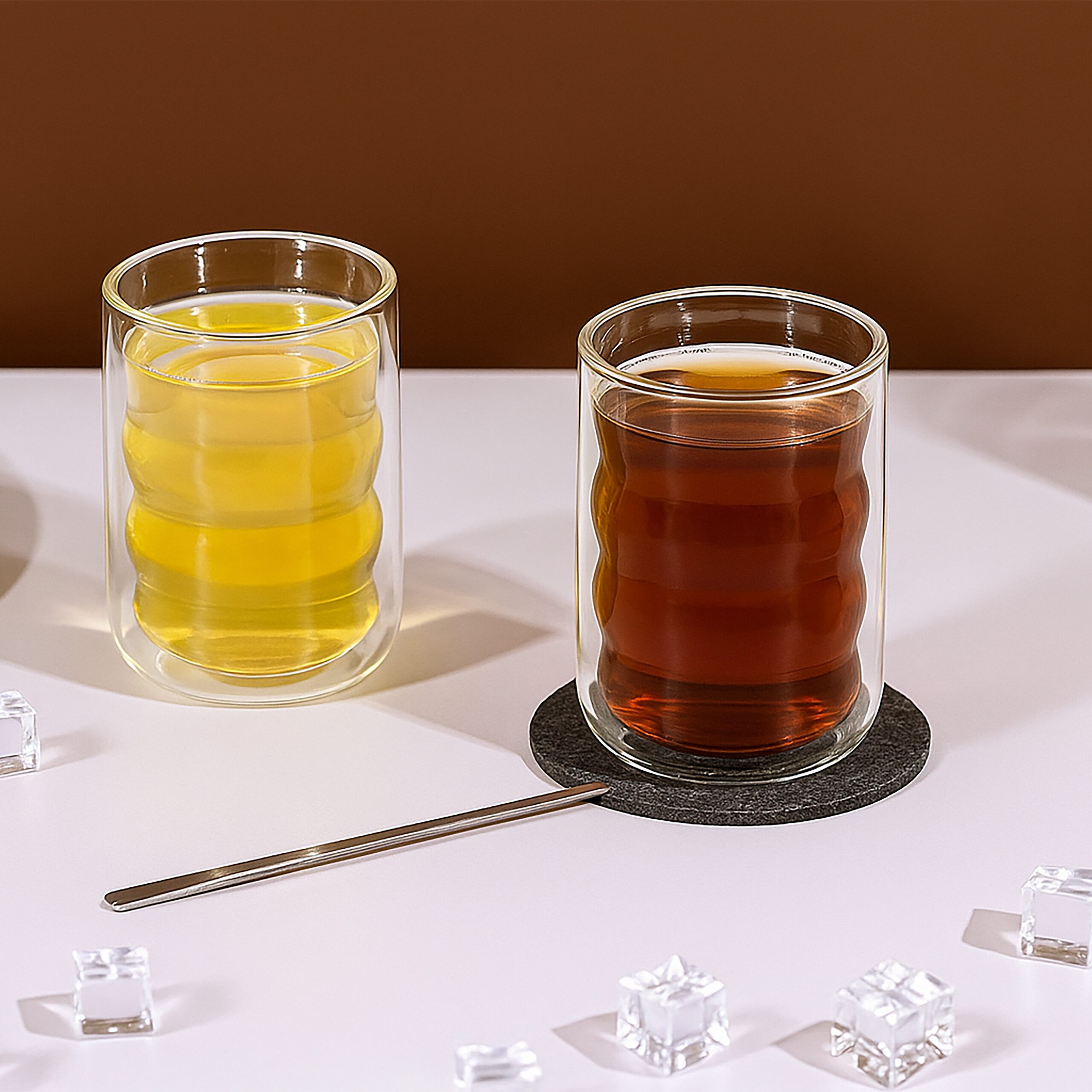 Zwei doppelwandige Gläser mit gelbem und braunem Getränk auf einem weißen Tisch, umgeben von Eiswürfeln.