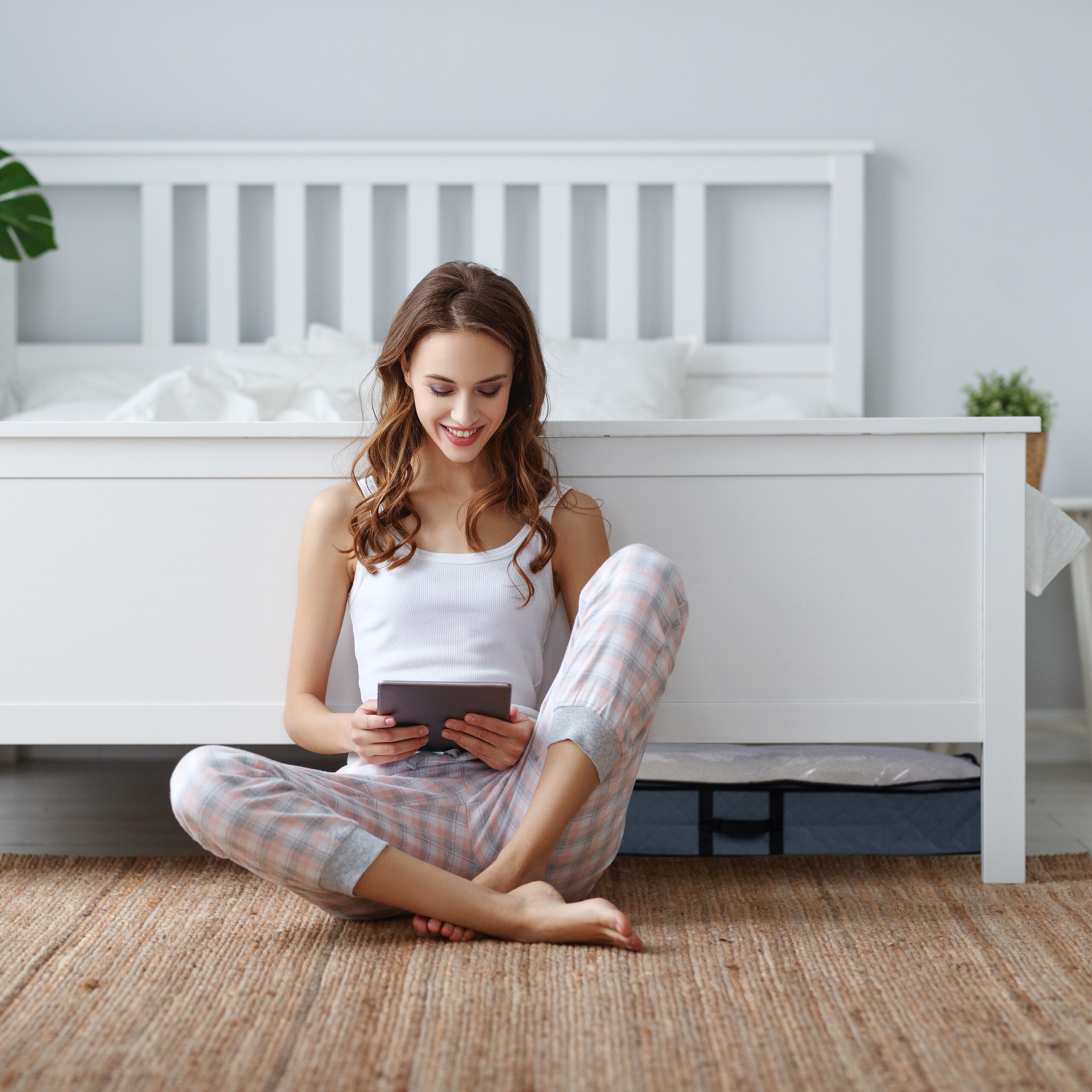 Eine lächelnde Frau im Schlafanzug sitzt vor einem weißen Bett auf einem Teppich und nutzt ein Tablet.