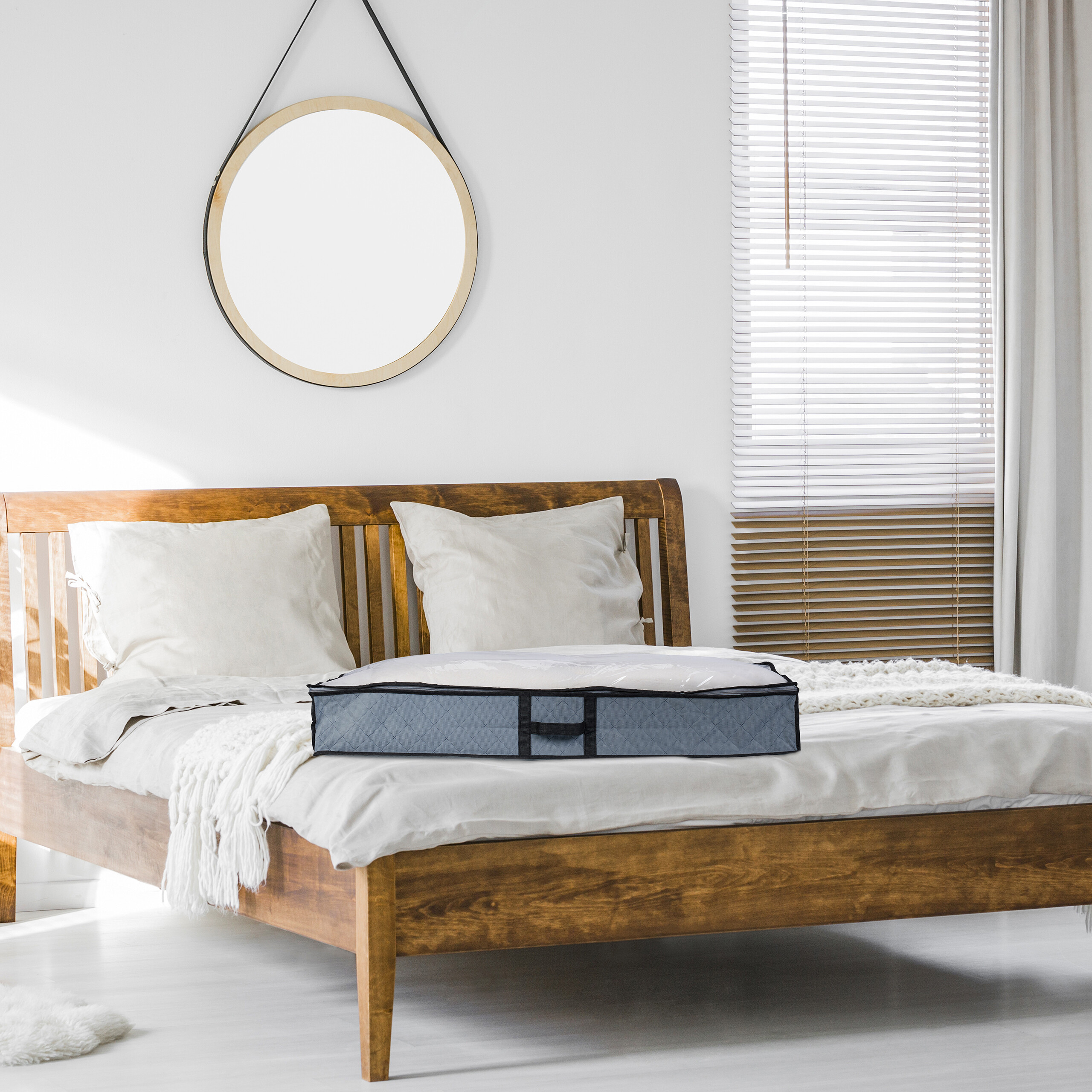 Ein Holzbett mit weißen Kissen und einer grauen Aufbewahrungstasche auf der Bettdecke in einem hellen Schlafzimmer.