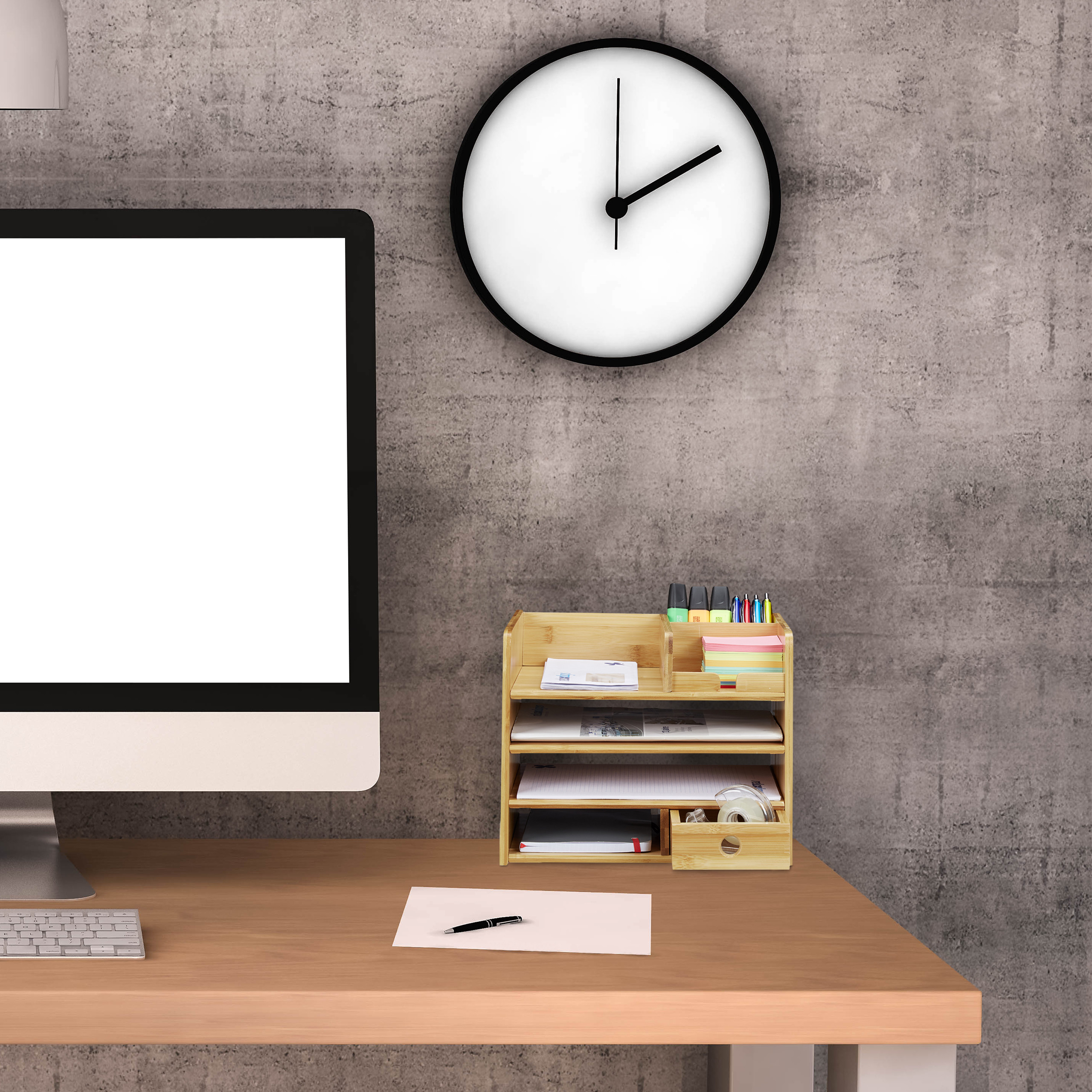 Büroarbeitsplatz mit Computer, Holzorganizer, Notizpapier und Wanduhr vor einer grauen Betonwand.