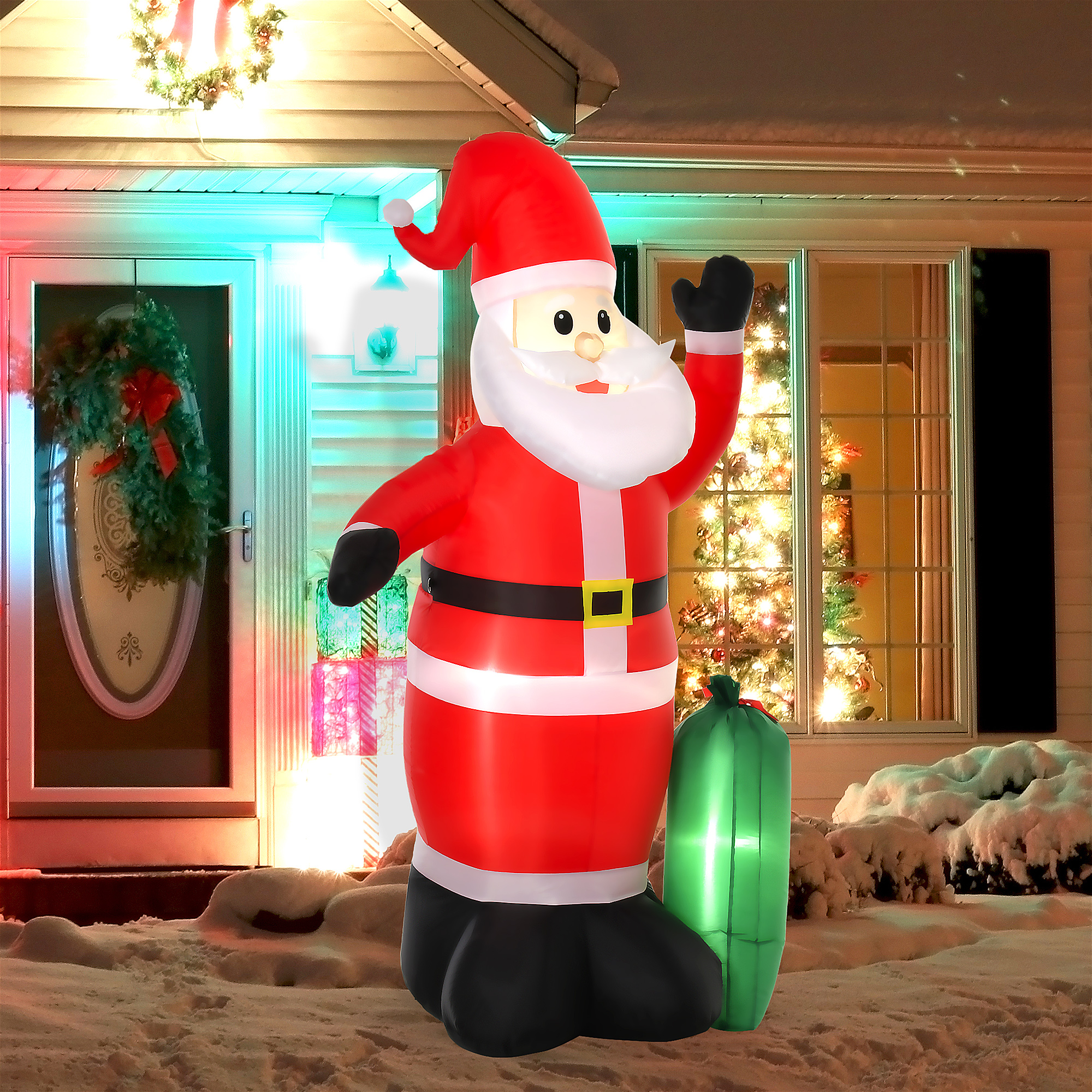 Un gran Papá Noel inflable está afuera de una casa cubierta de nieve por la noche, con un árbol de Navidad iluminado.