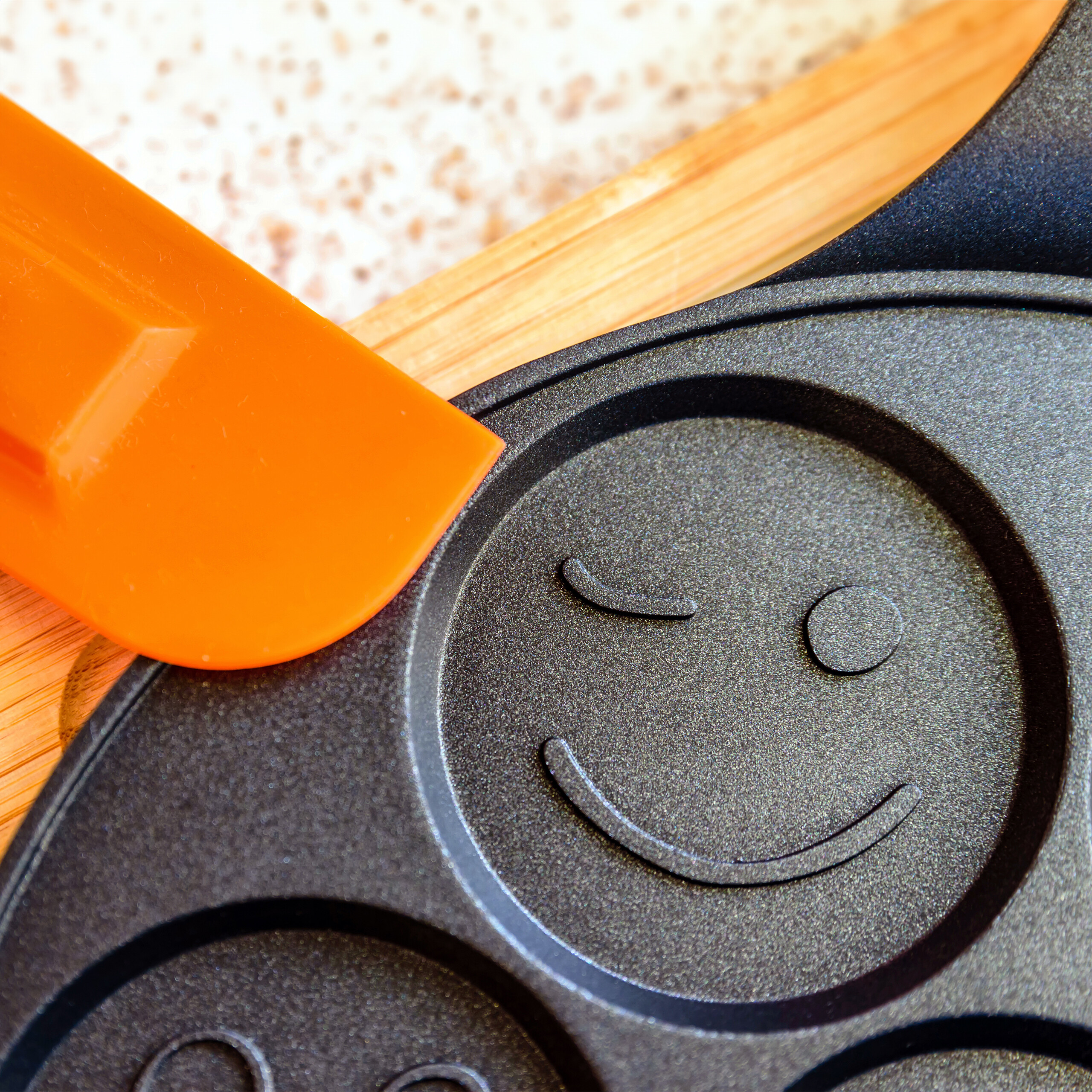 Eine schwarze Pfanne mit Winking-Smiley-Muster und ein orangefarbener Spatel auf Holz.