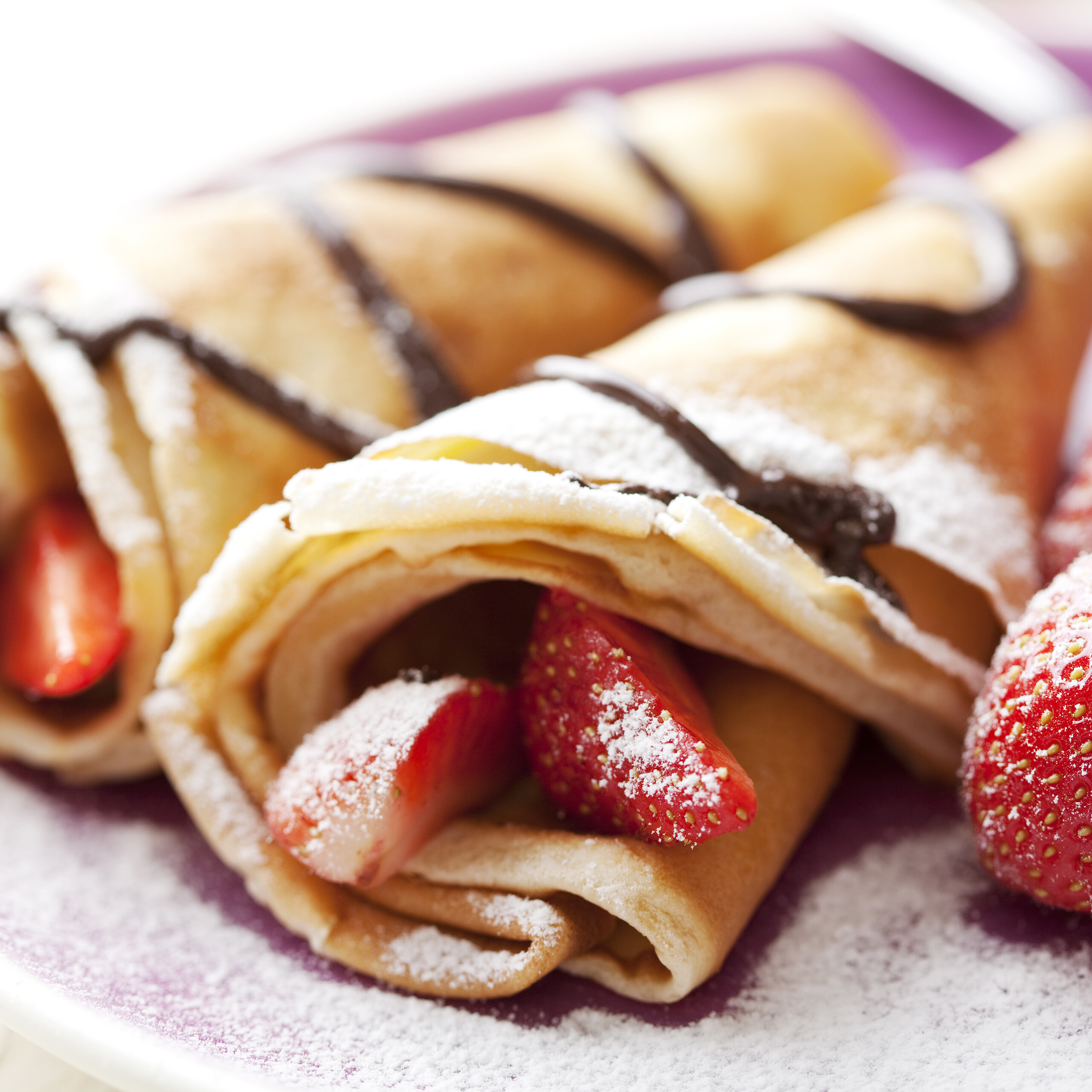 Gerollte Crêpes gefüllt mit Erdbeeren, bestäubt mit Puderzucker und dekoriert mit Schokoladensauce auf einem lila Teller.