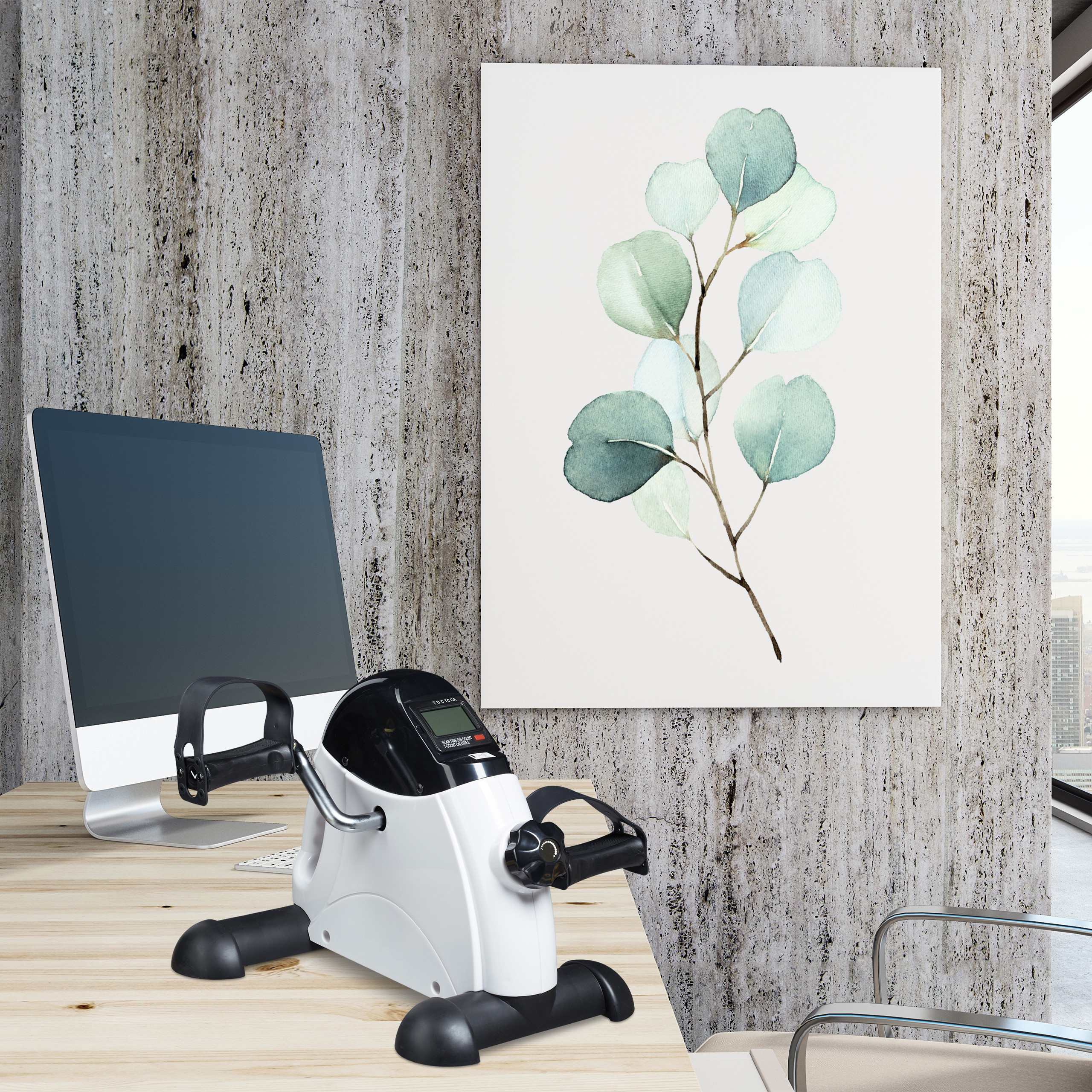 Heimtrainer auf einem Holztisch in einem Büro mit Computer und Pflanzenbild an der Wand.
