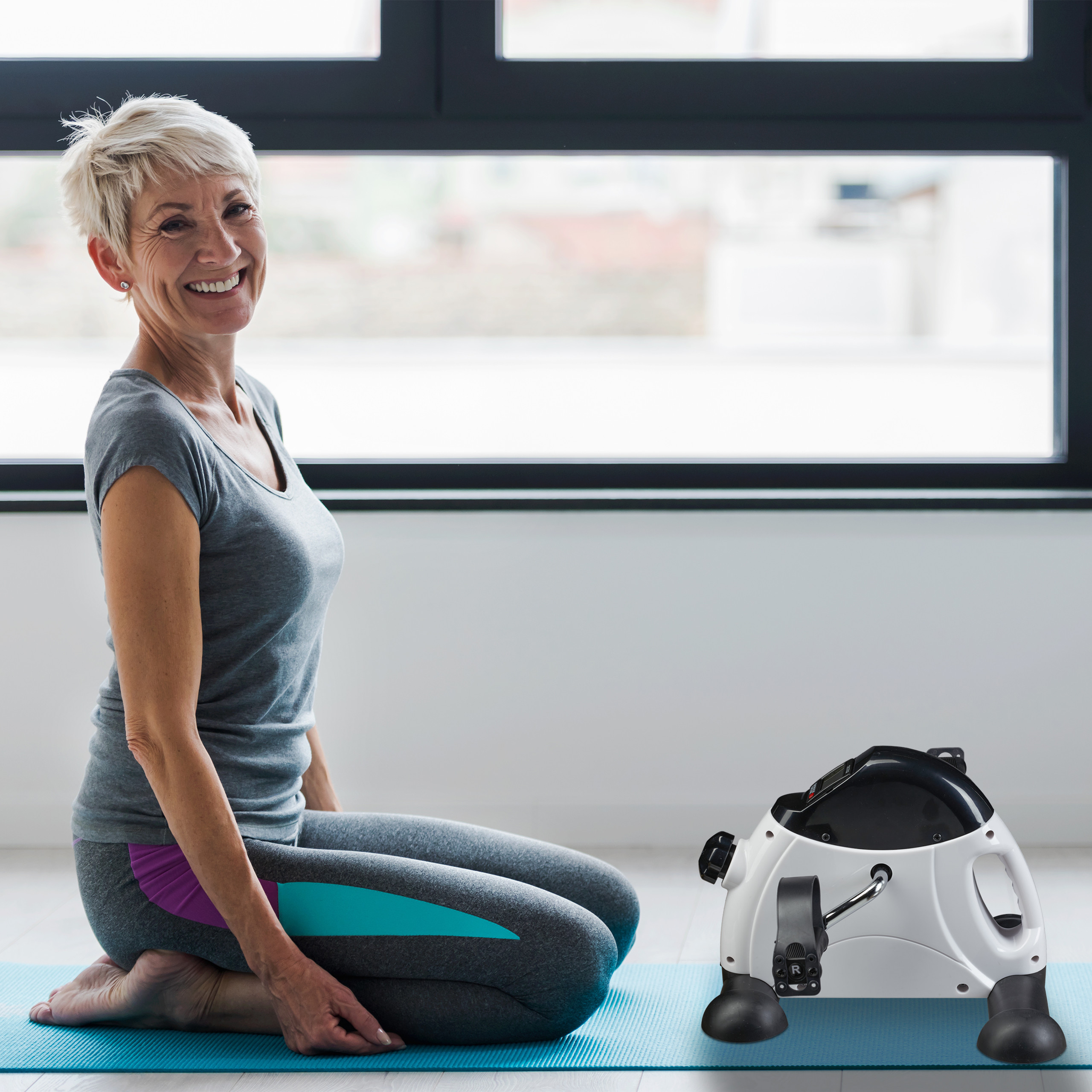 Ältere Frau mit kurzen blonden Haaren kniet auf einer blauen Yogamatte neben einem kleinen weißen Heimtrainer.