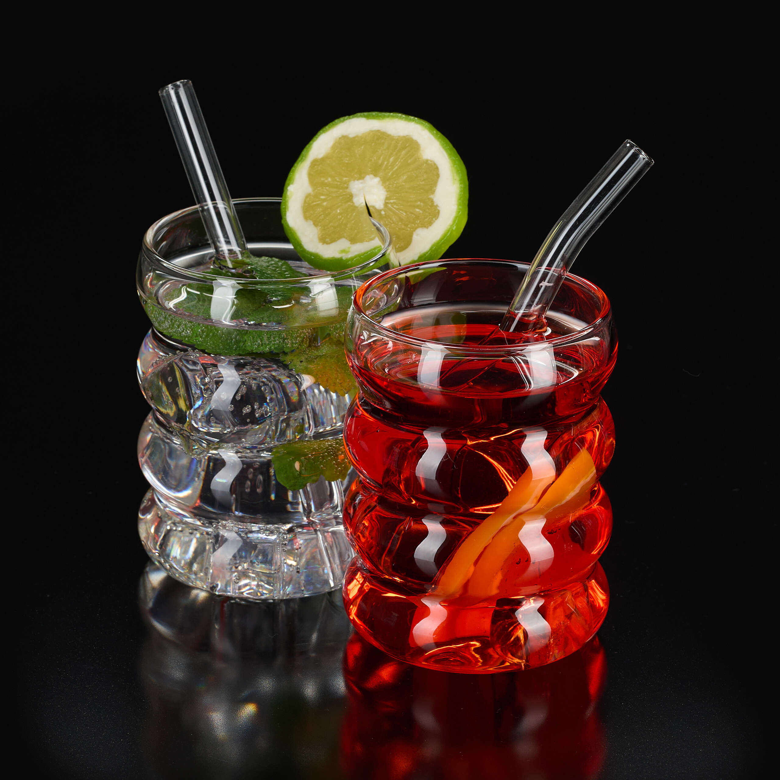 Zwei geriffelte Gläser mit Cocktails vor schwarzem Hintergrund. Links Mojito mit Limette, rechts ein roter Drink.