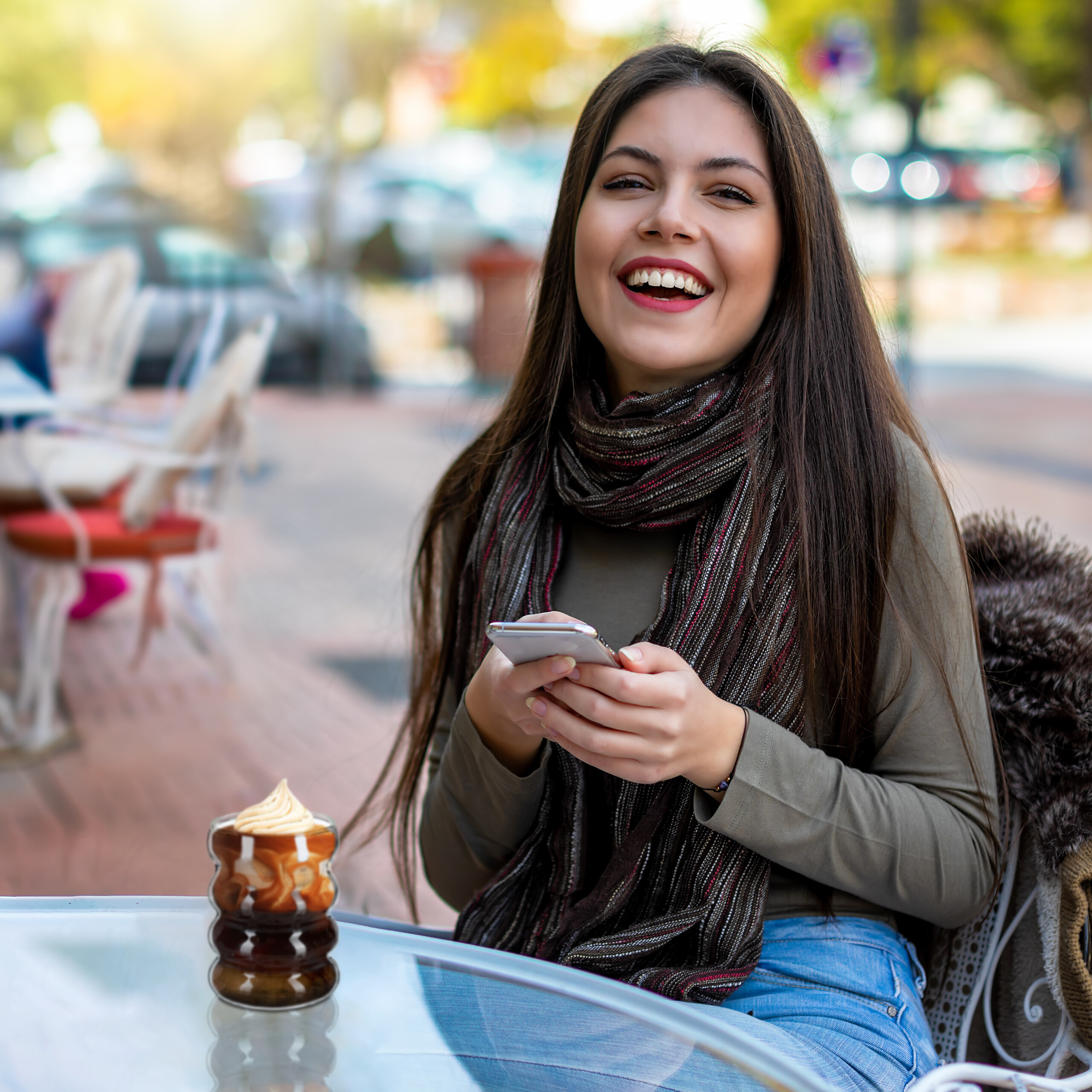 Eine lächelnde Frau sitzt im Café, hält ein Smartphone und hat einen Becher Kaffee vor sich auf dem Glastisch.