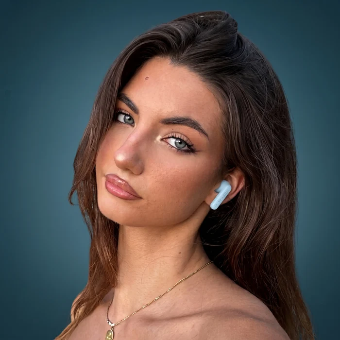 Una mujer castaña con un auricular azul y collar de oro sobre fondo oscuro.
