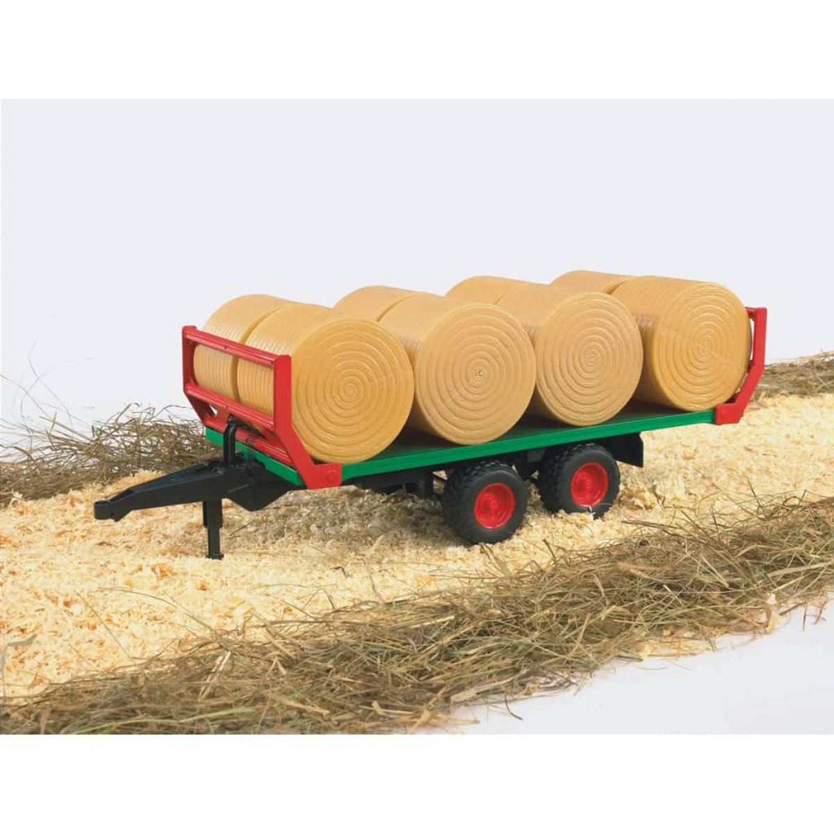 Ein grüner Anhänger mit rotem Rahmen transportiert gelbe Rundballen auf Holzspänen.