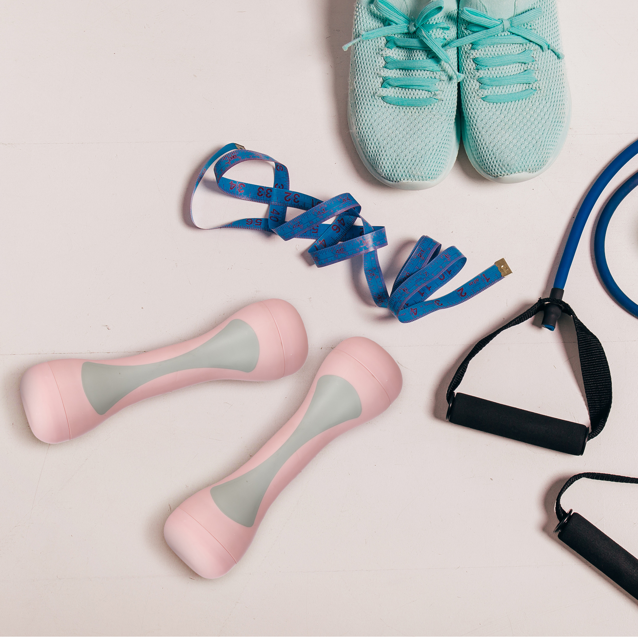 Fitnesszubehör mit rosafarbenen Hanteln, türkisfarbenen Schuhen, blauem Maßband und Widerstandsband auf weißem Hintergrund.