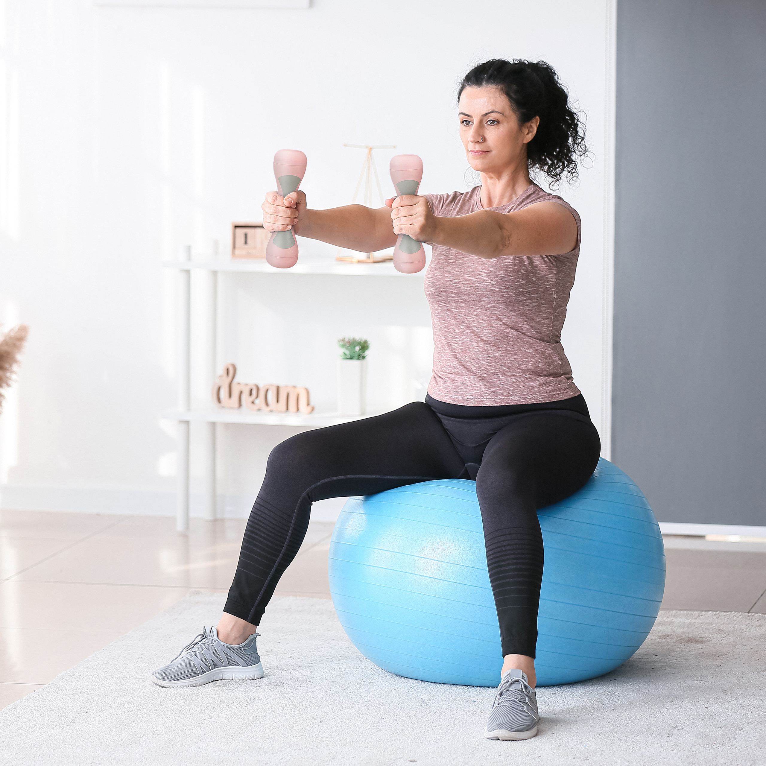 Eine Frau sitzt auf einem blauen Gymnastikball, hält rosa Hanteln und hebt die Arme vor.