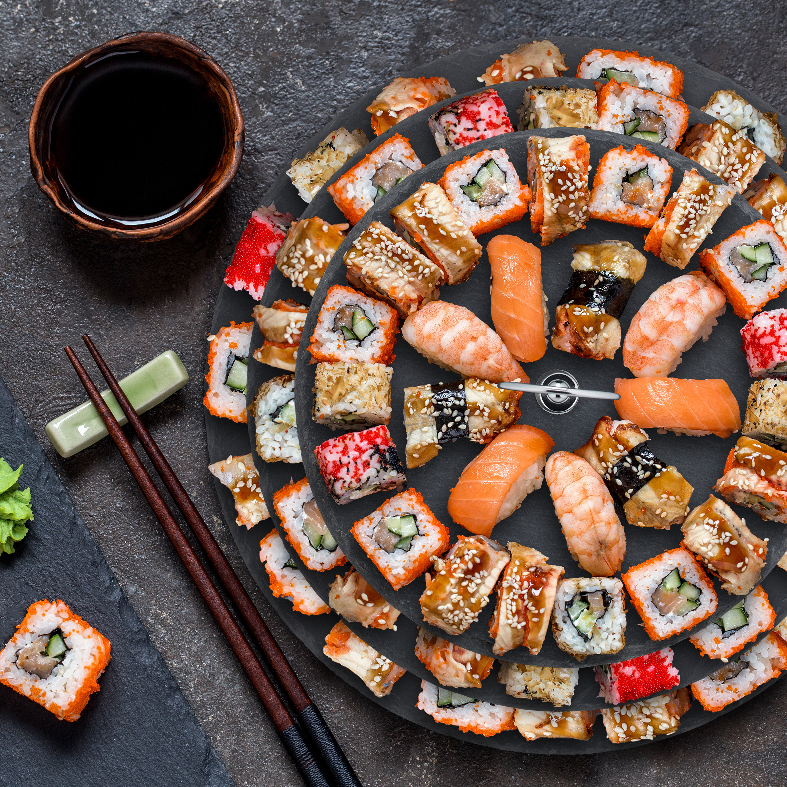 Verschiedene Sushi-Rollen und Nigiri auf einer dreistöckigen Schieferplatte mit Sojasauce und Stäbchen.
