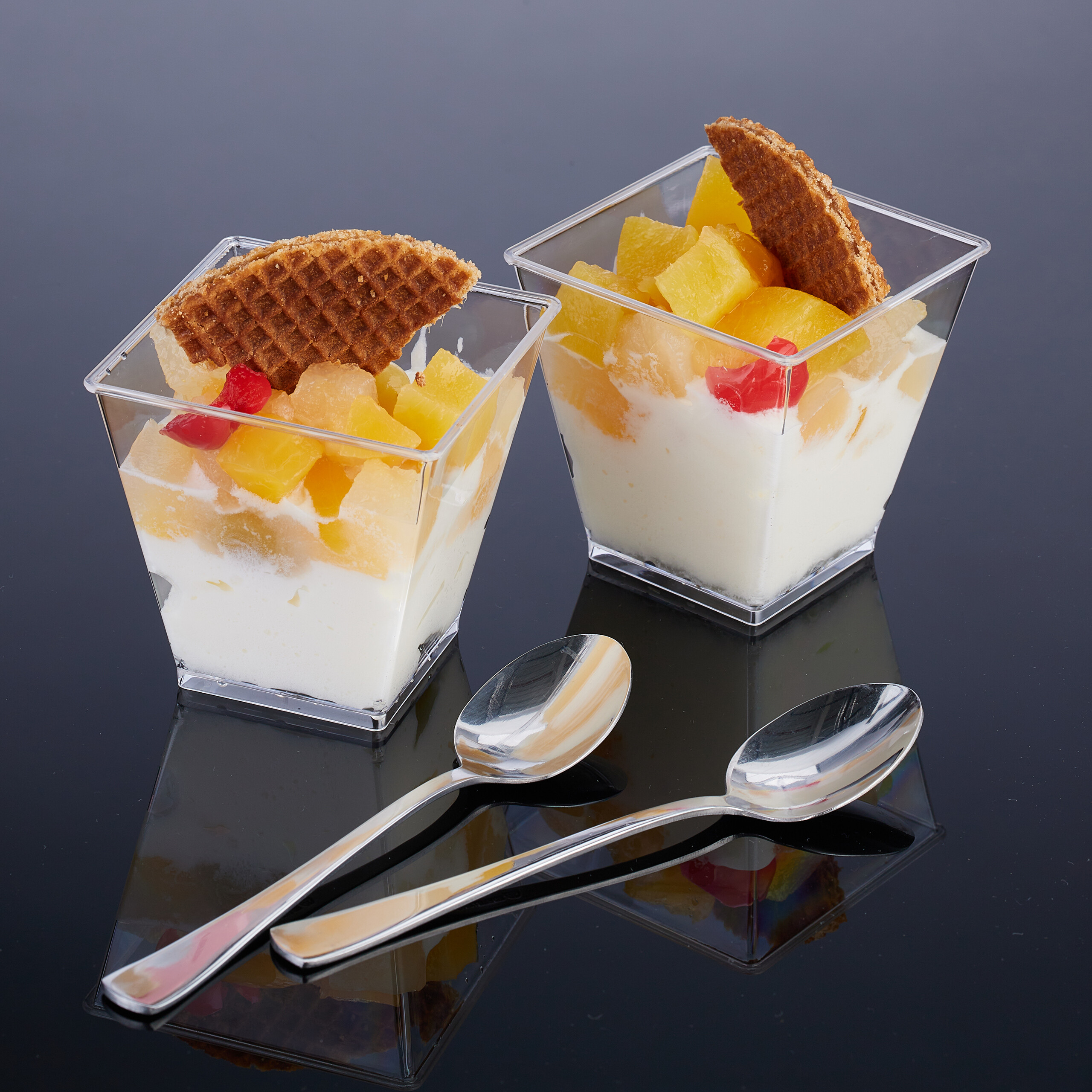 Zwei Dessertgläser mit Joghurt, gelben Früchten und Waffelkeksen stehen auf glänzender Fläche mit zwei Silberlöffeln.