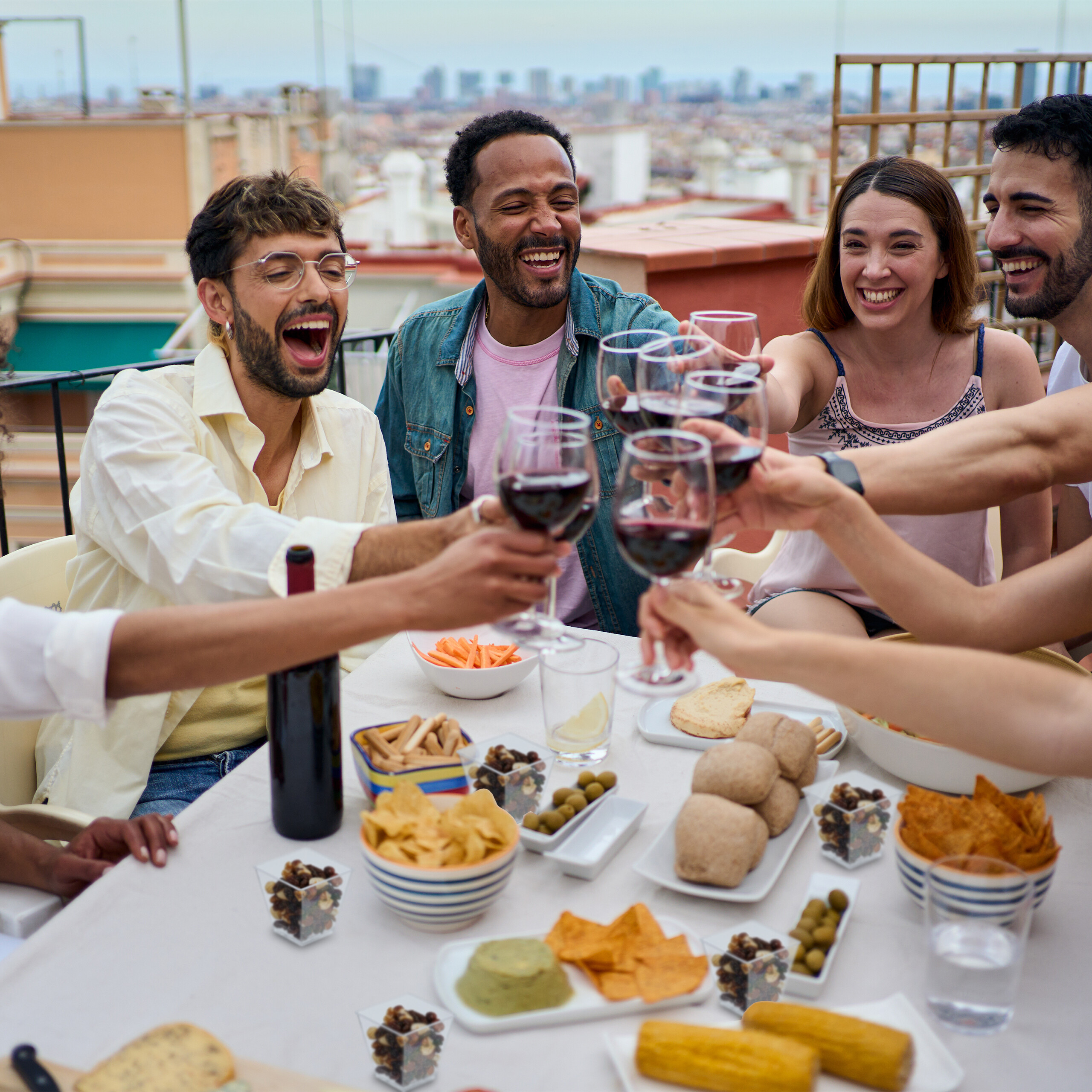 Freunde stoßen auf einer sonnigen Dachterrasse mit Rotwein und Snacks an.