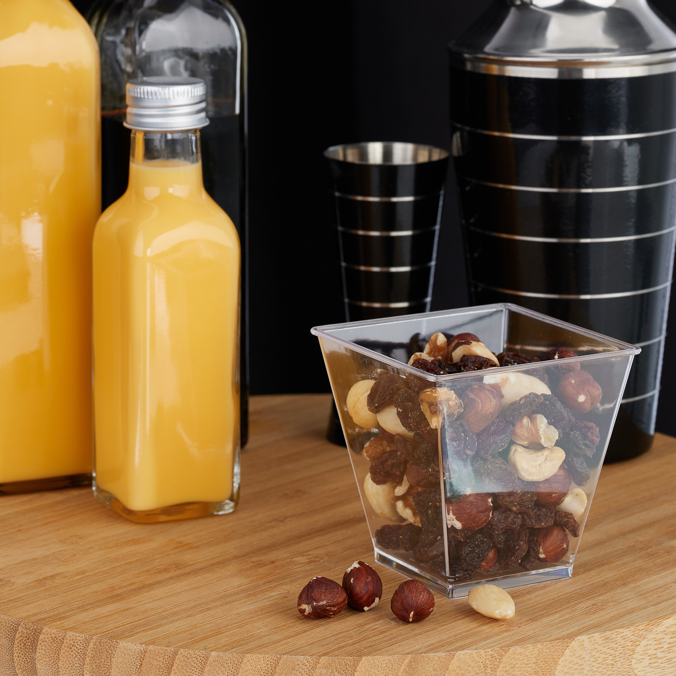 Nüsse und Rosinen in einem transparenten Becher auf einem Holztisch, umgeben von Flaschen und einem Cocktailshaker.