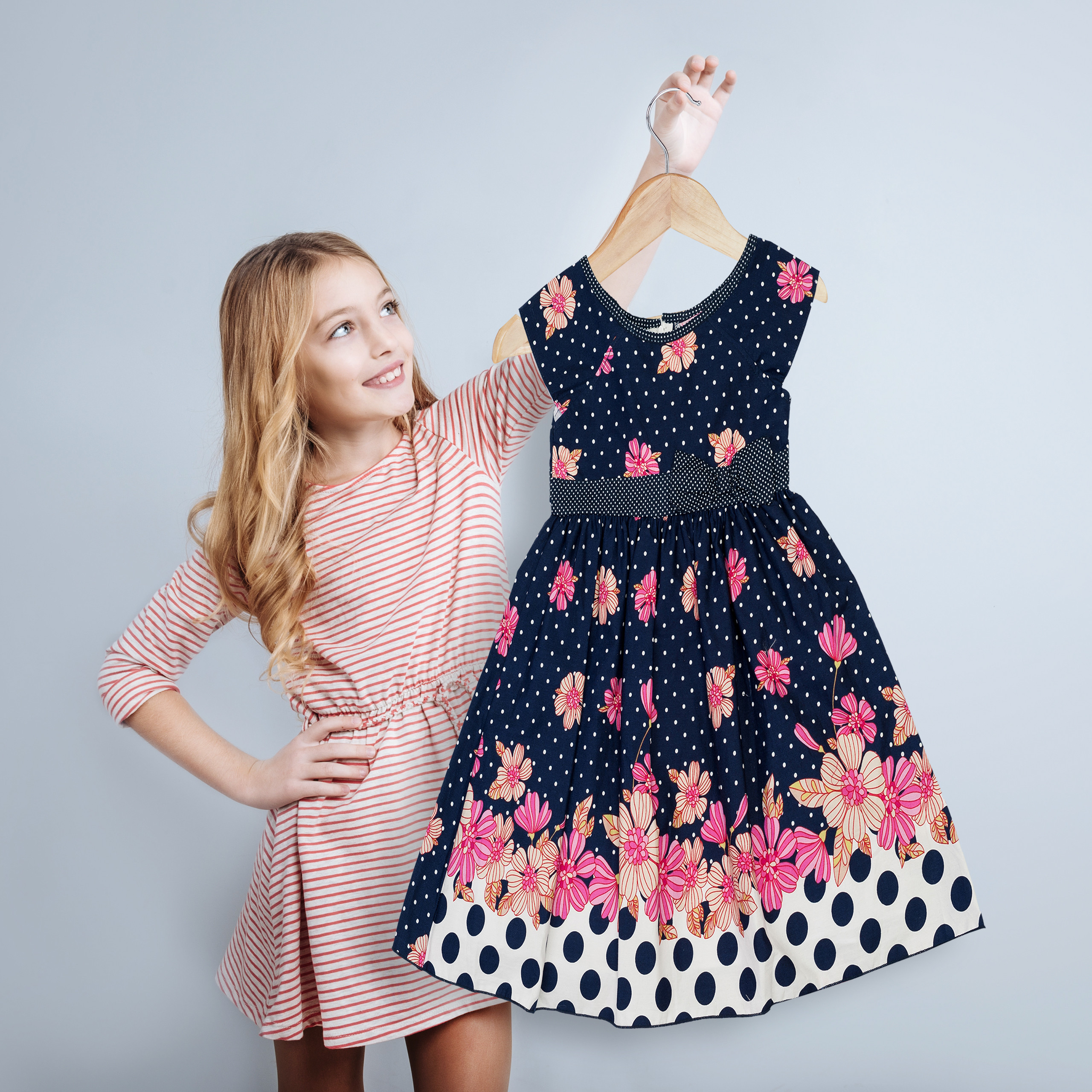 Ein blondes Mädchen in einem gestreiften Kleid hält ein blaues Sommerkleid mit Blumenmuster an einem Bügel hoch.