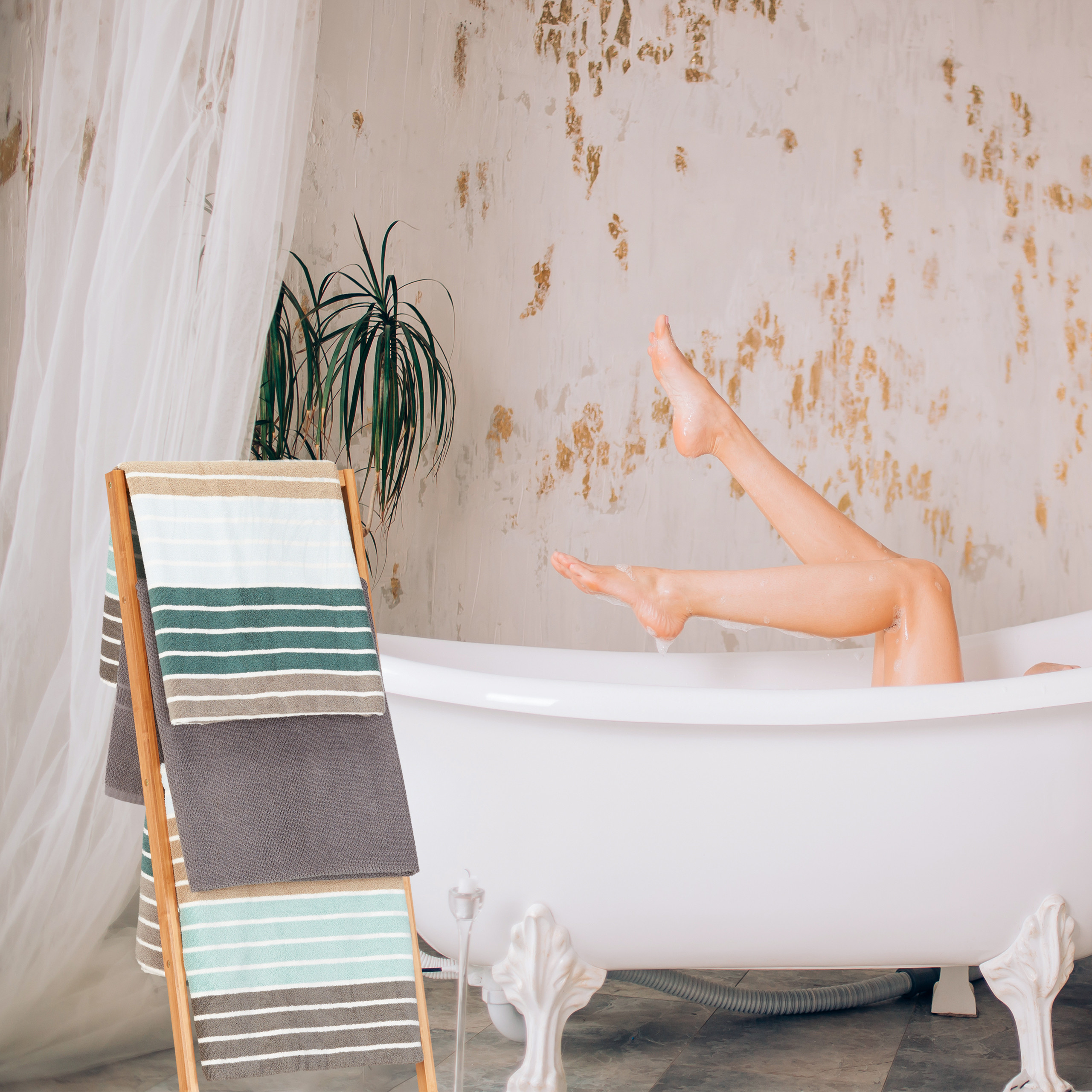 Beine einer Person ragen aus einer weißen Badewanne. Links steht ein Holzwäscheständer mit bunten Handtüchern.