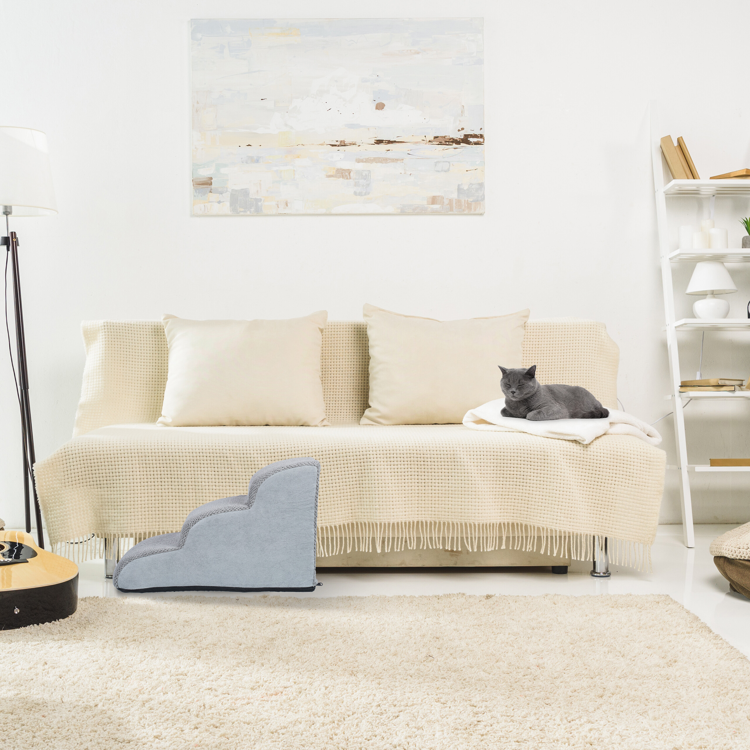 Eine graue Katze ruht auf einer beigen Couch mit Kissen und Decke in einem hellen Wohnzimmer.