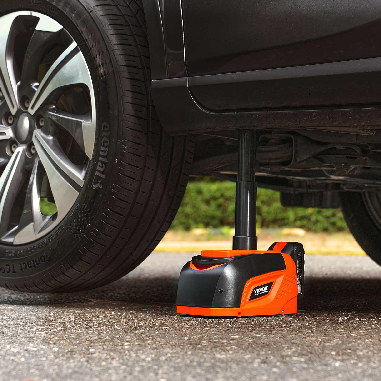 Un gato hidráulico eléctrico en negro y naranja levanta el chasis de un coche negro en el suelo pavimentado.