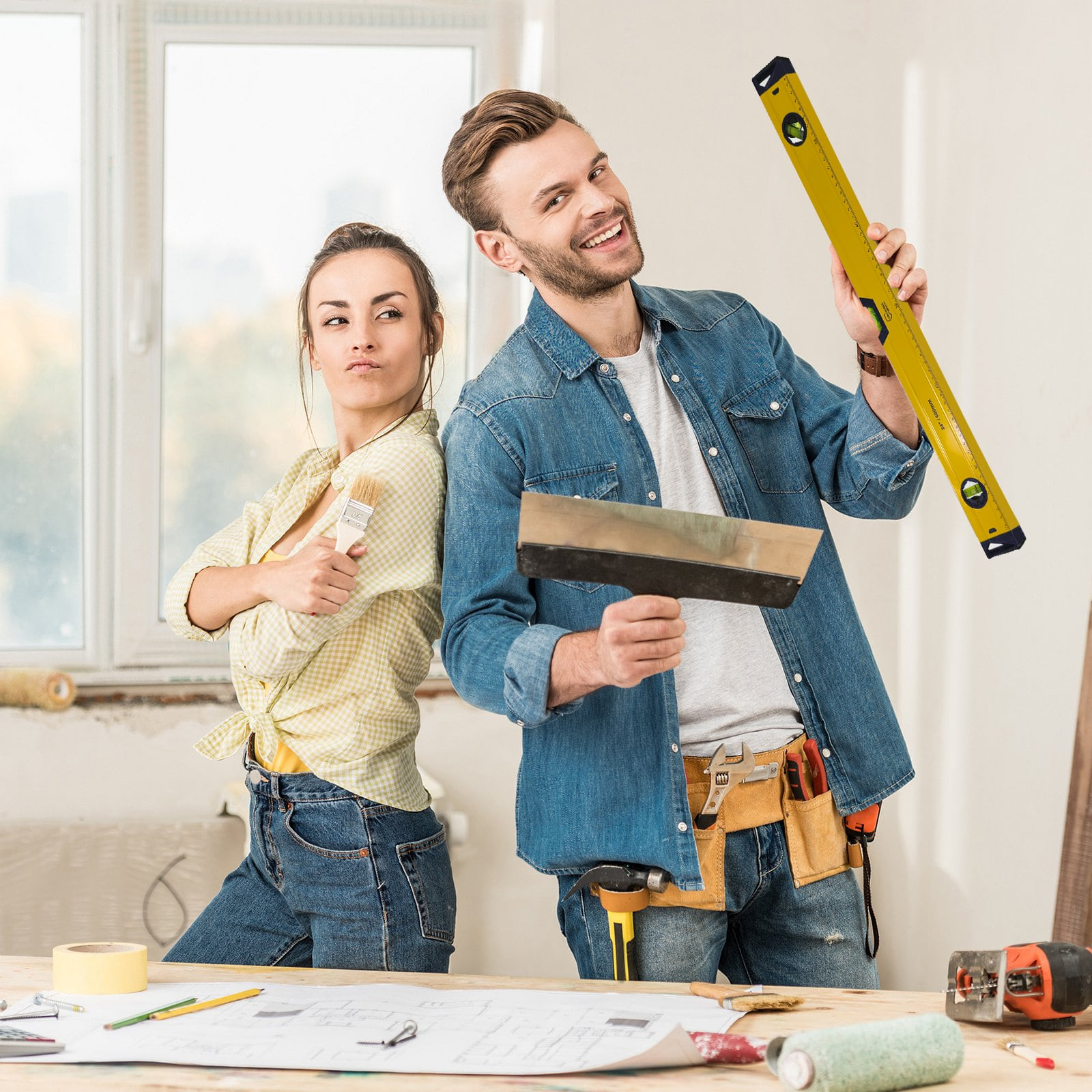 Ein Handwerker und eine Handwerkerin posieren lächelnd mit Werkzeugen vor einem renovierungsbedürftigen Raum.