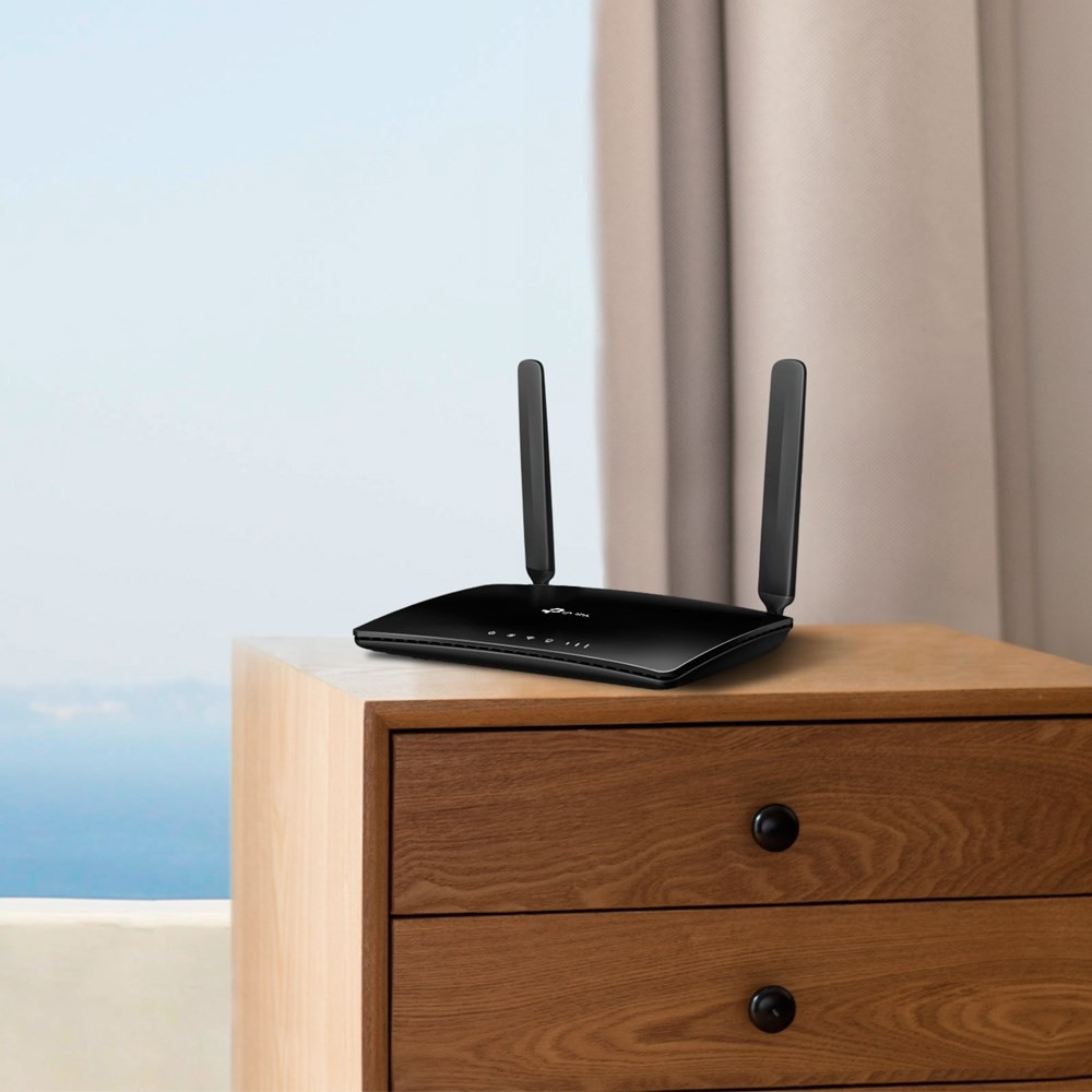 Ein schwarzer WLAN-Router mit zwei Antennen steht auf einer Holzkommode vor einem hellen Fenster und Vorhang.