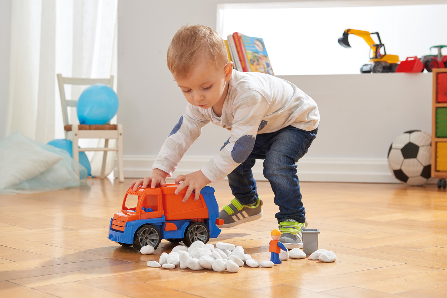 Niño pequeño jugando en suelo de madera con un camión de basura de juguete naranja y azul y piedras blancas.