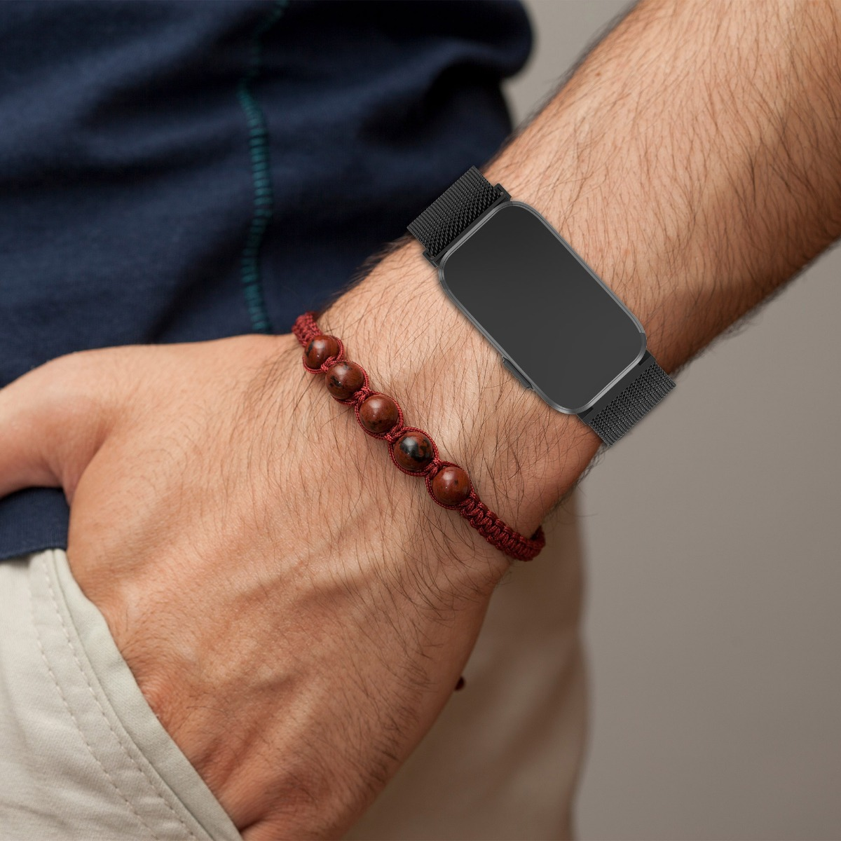 Eine Person trägt eine schwarze Smartwatch mit Mesh-Armband und ein rotes Perlenarmband am Handgelenk.
