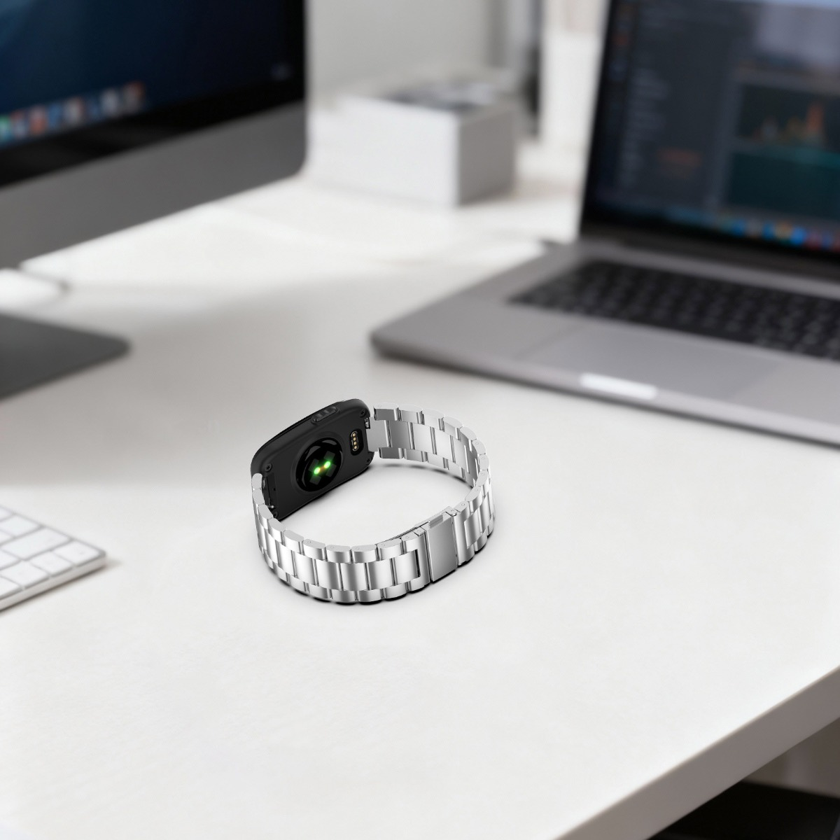 Eine Smartwatch mit silbernem Metallarmband liegt auf einem weißen Schreibtisch mit Laptop und Monitor im Hintergrund.