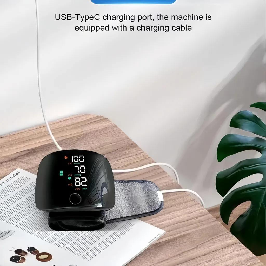 Ein schwarzes Blutdruckmessgerät mit digitaler Anzeige und Ladekabel auf einem Holztisch, daneben eine Pflanze.