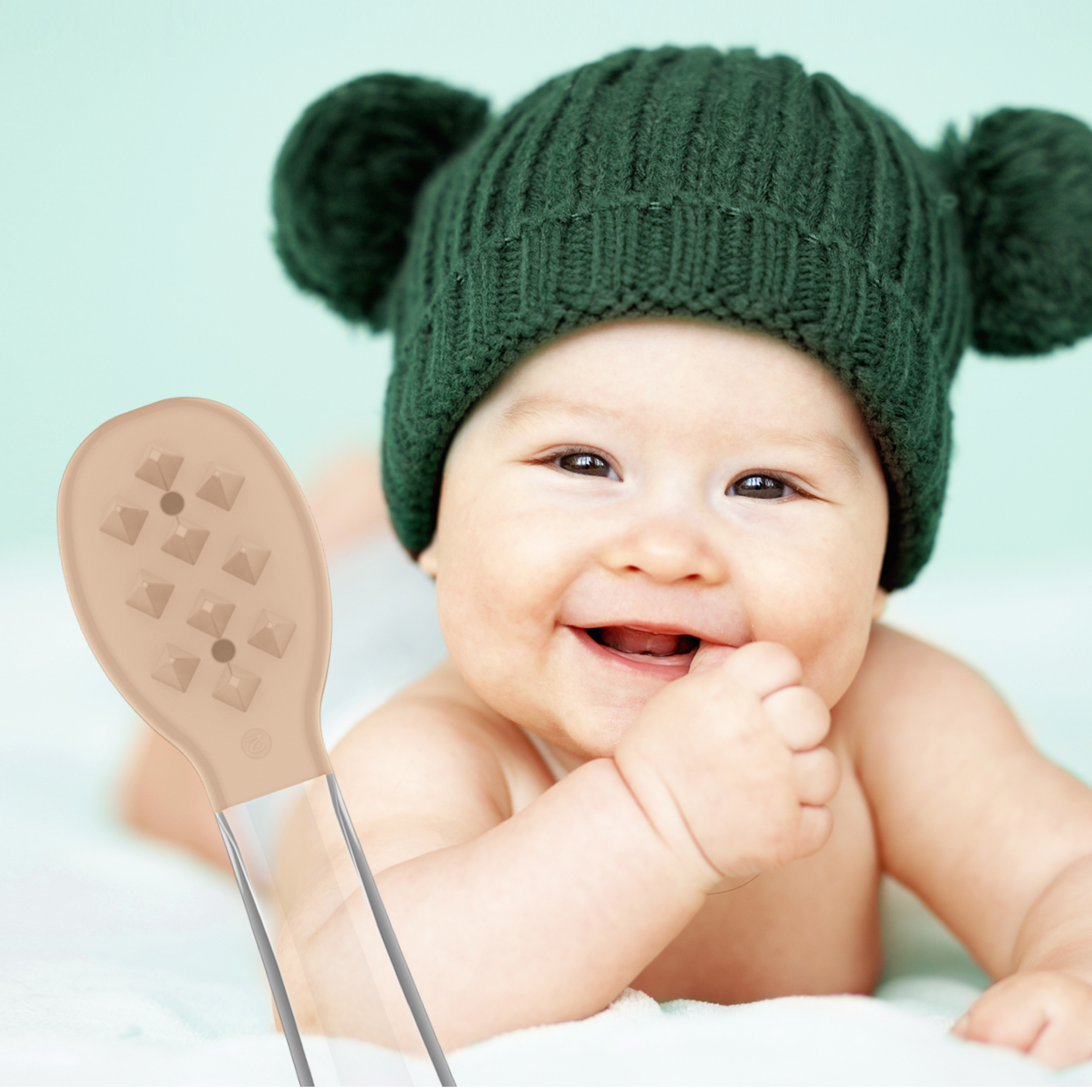 Ein lächelndes Baby mit einer grünen Mütze und Pompons liegt neben einem beigen Silikon-Babylöffel.