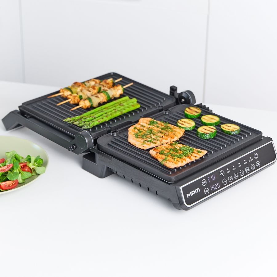Otwarty grill elektryczny na białym blacie piecze szaszłyki, szparagi, rybę i plastry cukinii, obok stoi sałatka.