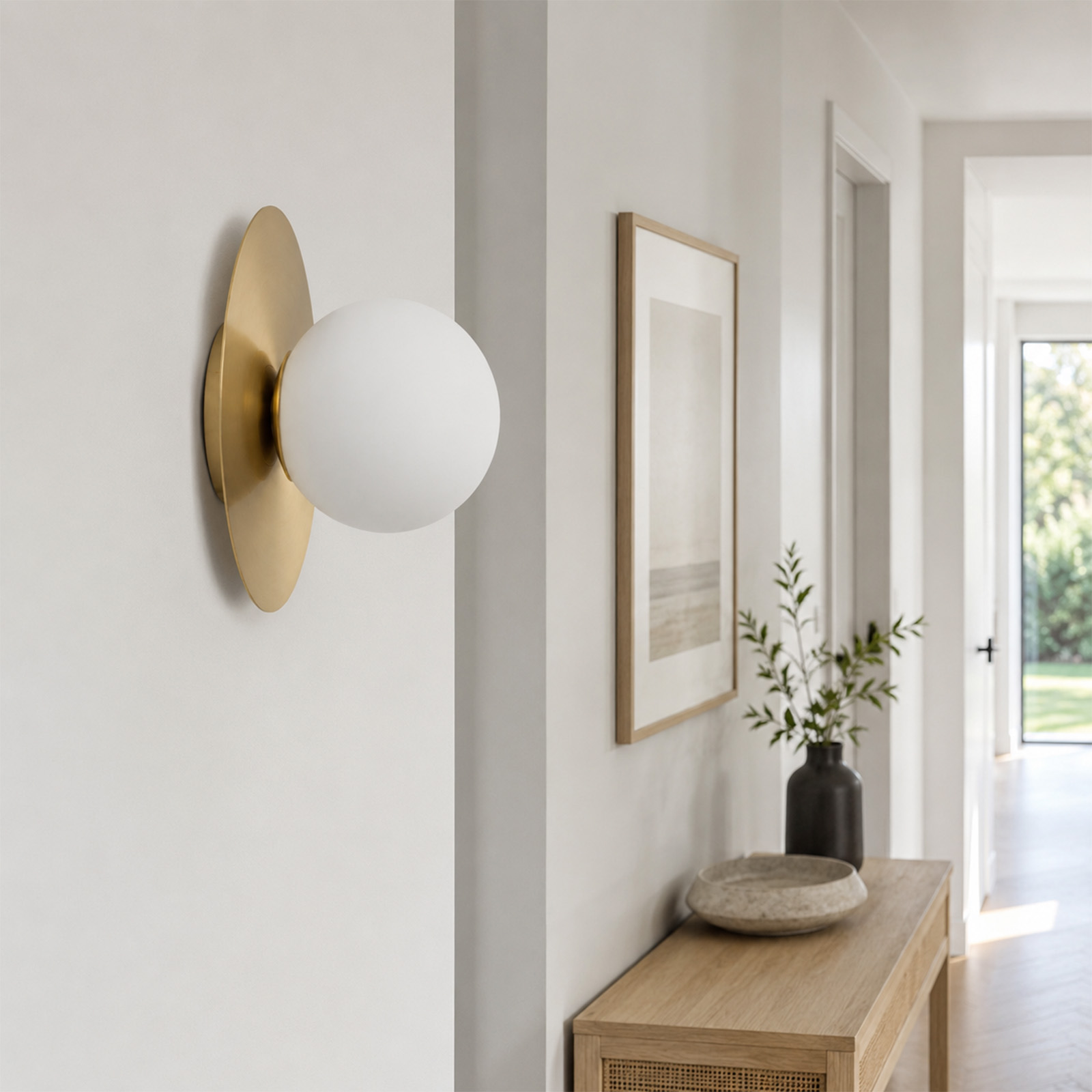 Una lámpara de pared blanca y redonda con soporte dorado en un pasillo luminoso con mesa de consola y cuadro.
