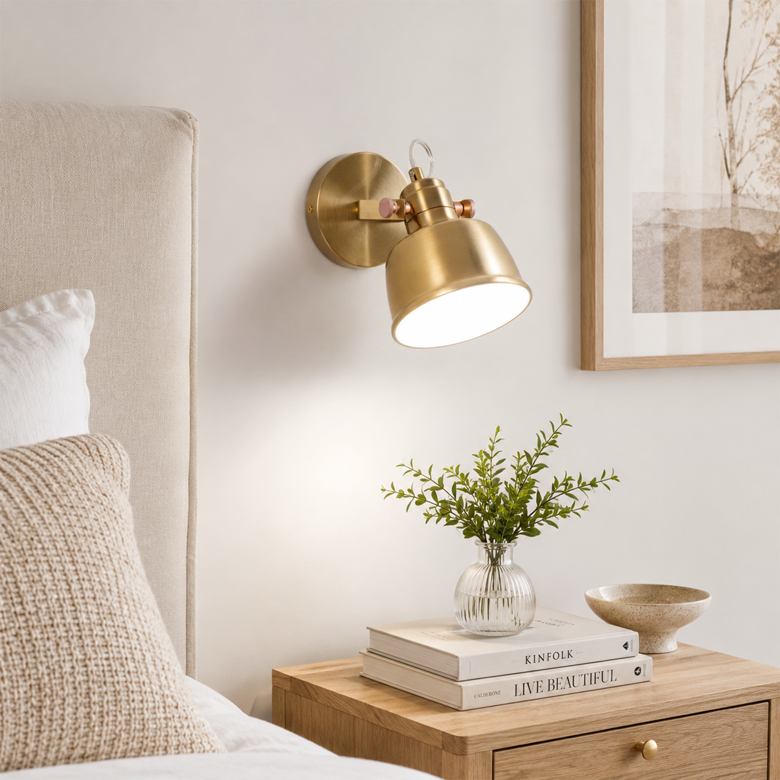 Una lámpara de pared dorada ilumina una mesita de noche con libros y una planta junto a una cama.
