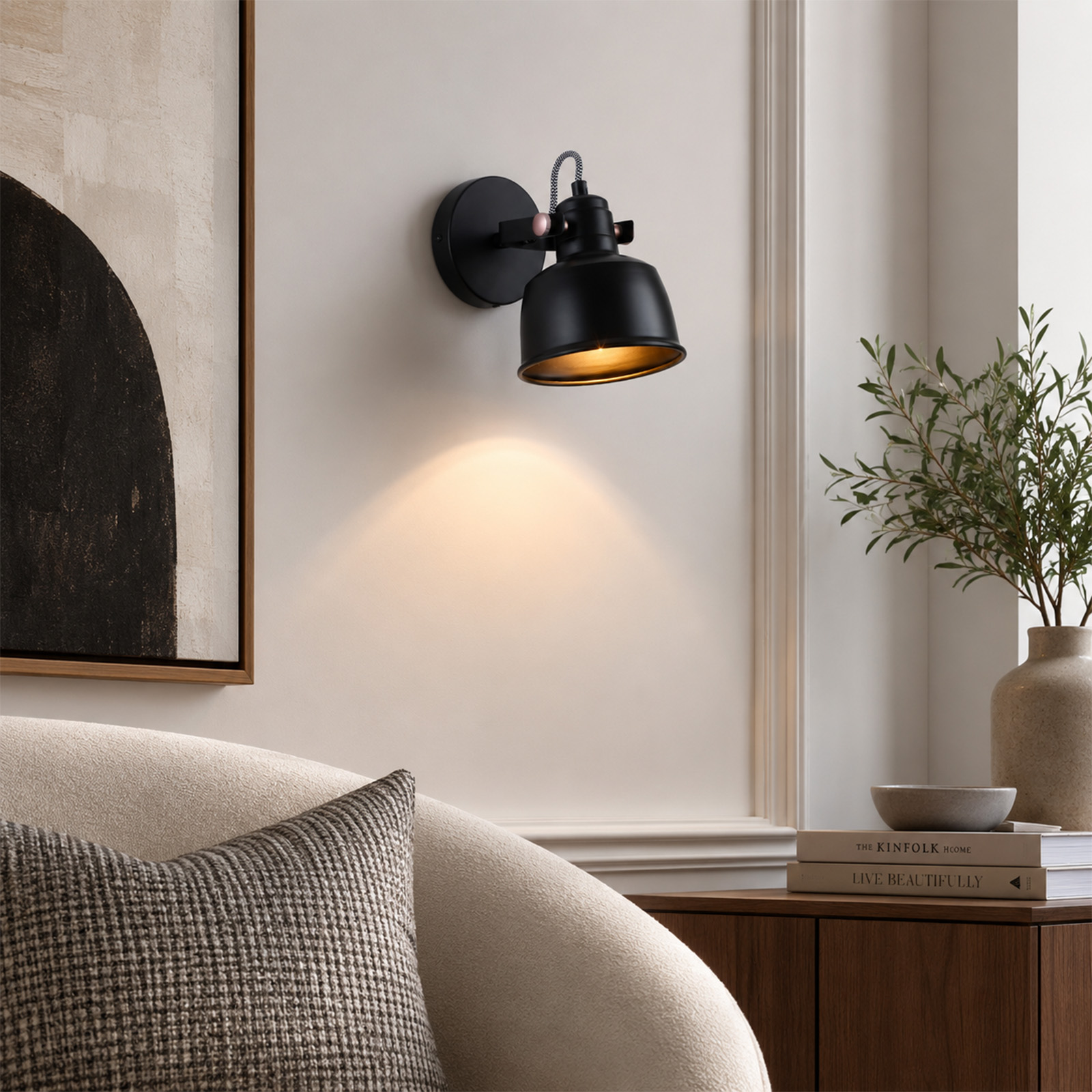 Una lámpara de pared negra brilla en una pared blanca sobre un sillón blanco con un cojín estampado.