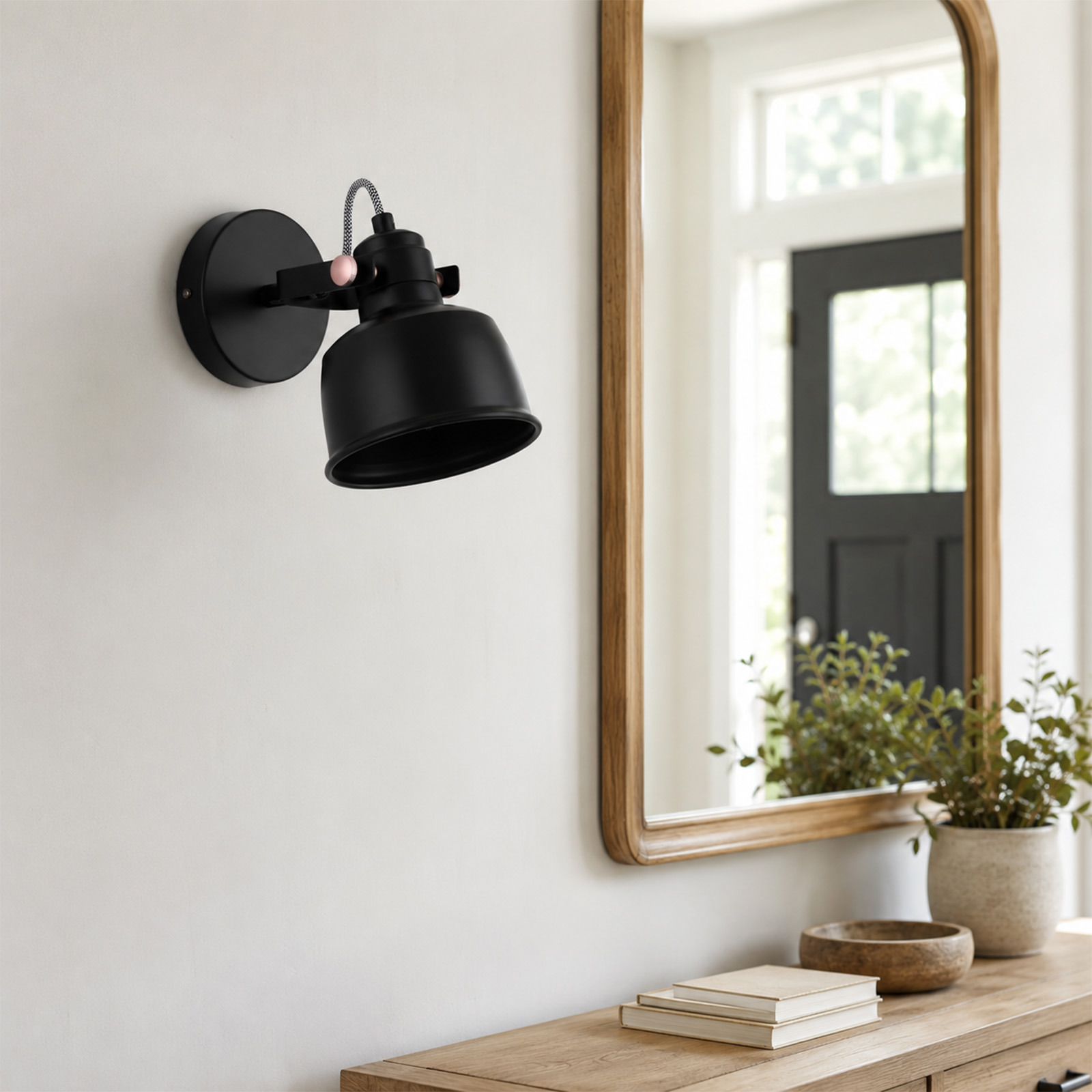 Un aplique de pared negro en una pared blanca sobre una mesa consola de madera con espejo y planta.