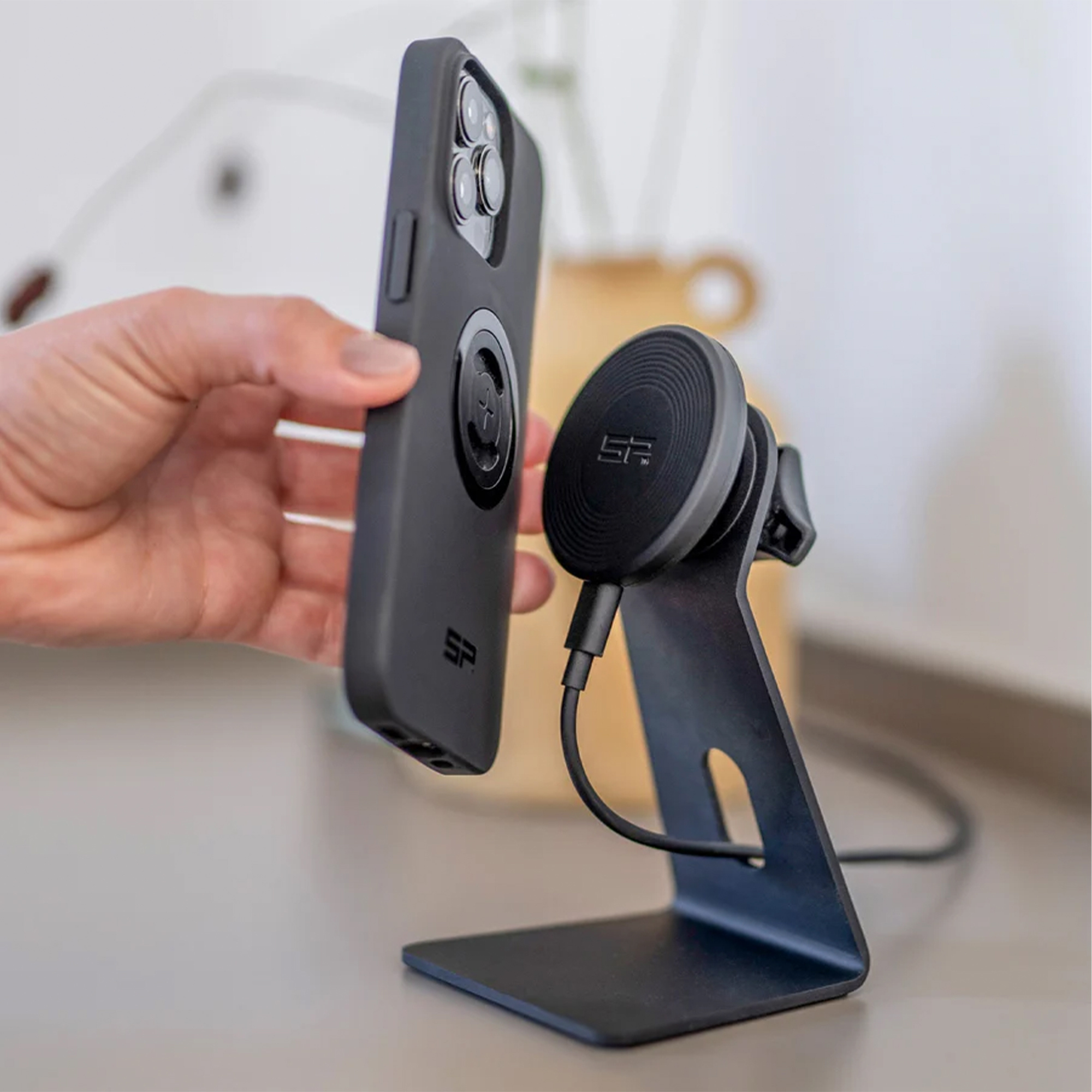 Eine Hand hält ein schwarzes Smartphone mit Hülle nahe an eine magnetische Tischhalterung mit Ladekabel.