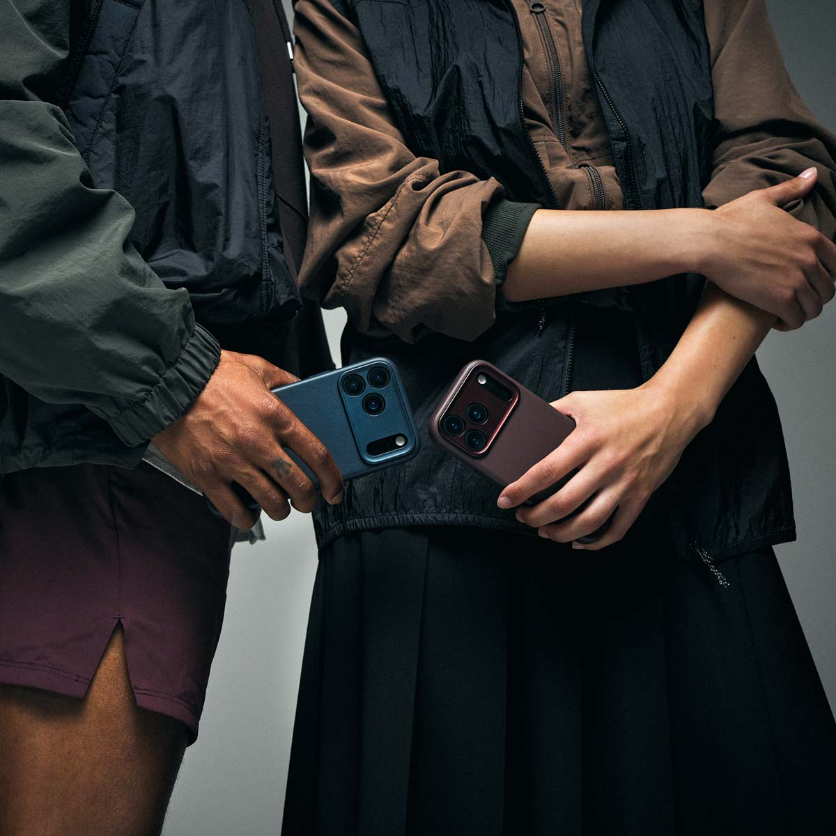 Zwei Personen in dunklen Jacken halten Smartphones mit bunten Hüllen vor grauem Hintergrund.