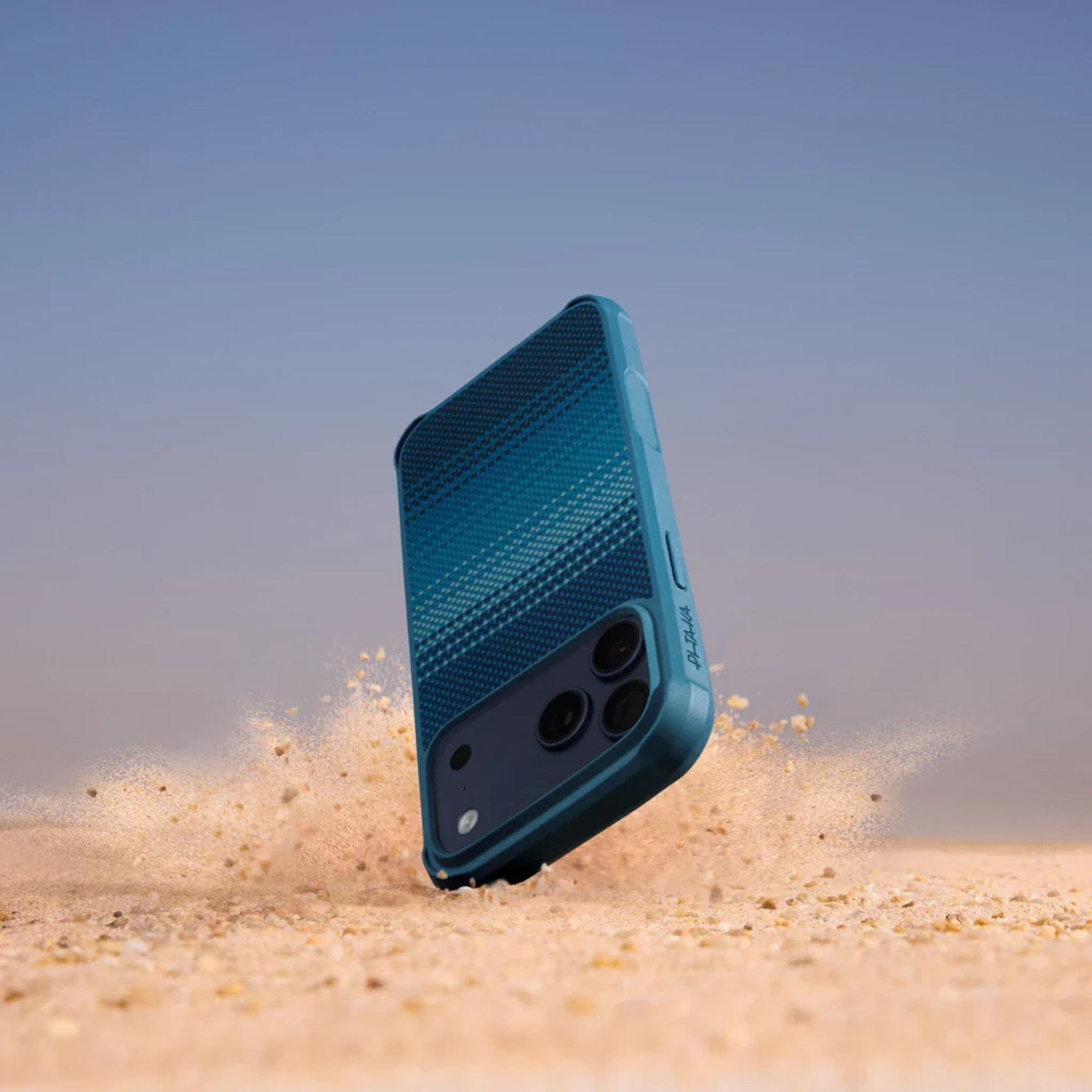 Ein blaues Smartphone mit strukturierter Hülle trifft auf Sand und wirbelt Staub auf.