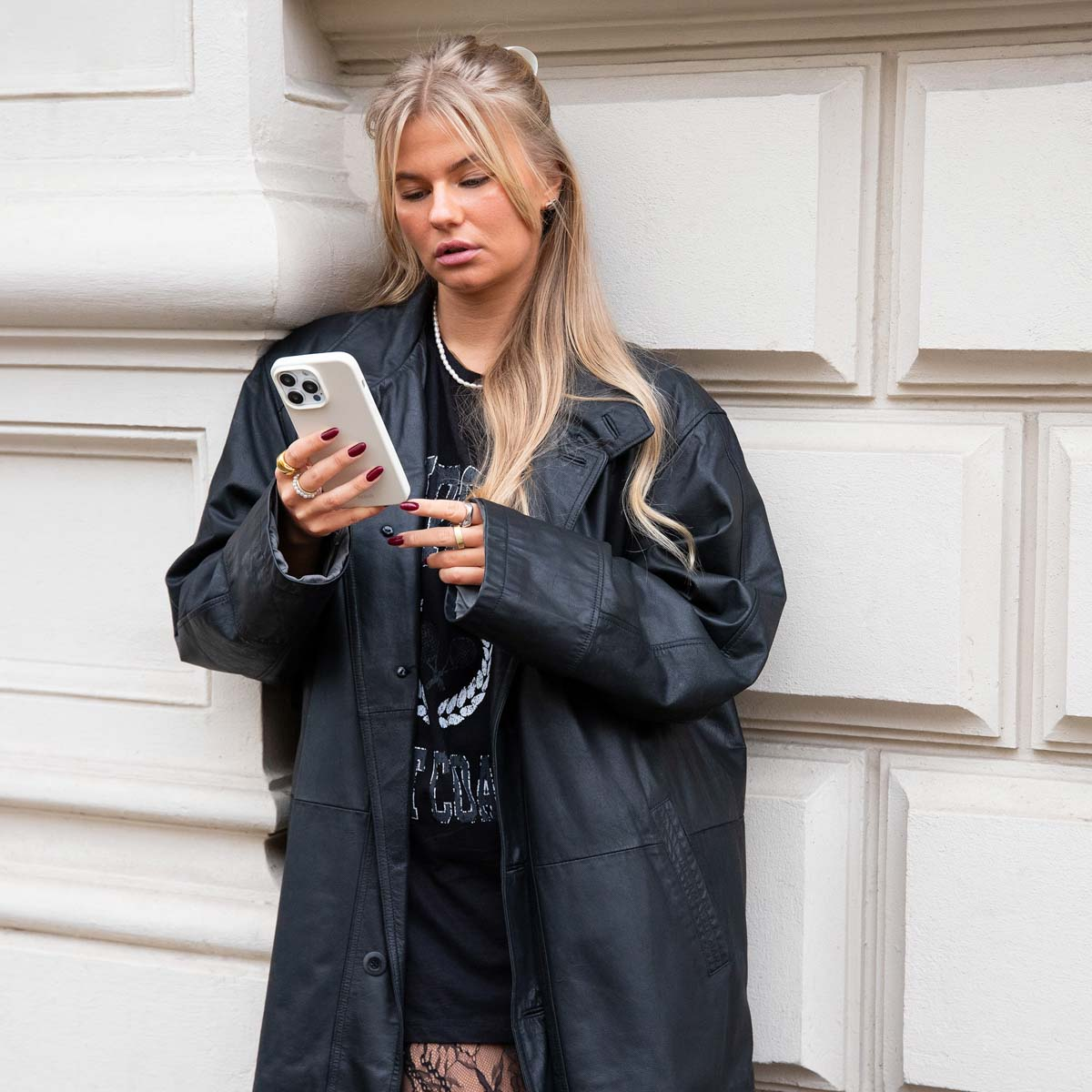 Eine blonde Frau in schwarzem Ledermantel und T-Shirt lehnt an einer Wand und blickt auf ihr weißes Smartphone.