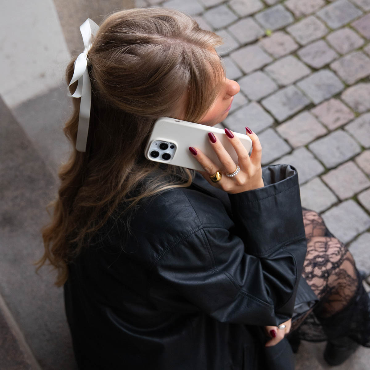 Eine blonde Frau mit einer weißen Schleife im Haar telefoniert auf einem Kopfsteinpflaster.