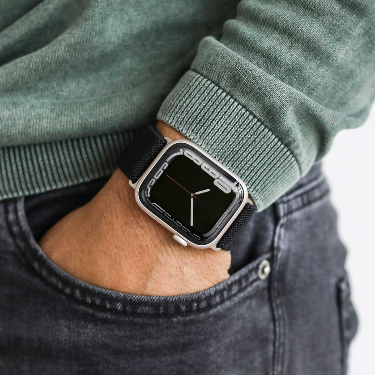 Osoba nosząca srebrny smartwatch z czarnym paskiem, zielonym swetrem i szarymi dżinsami.
