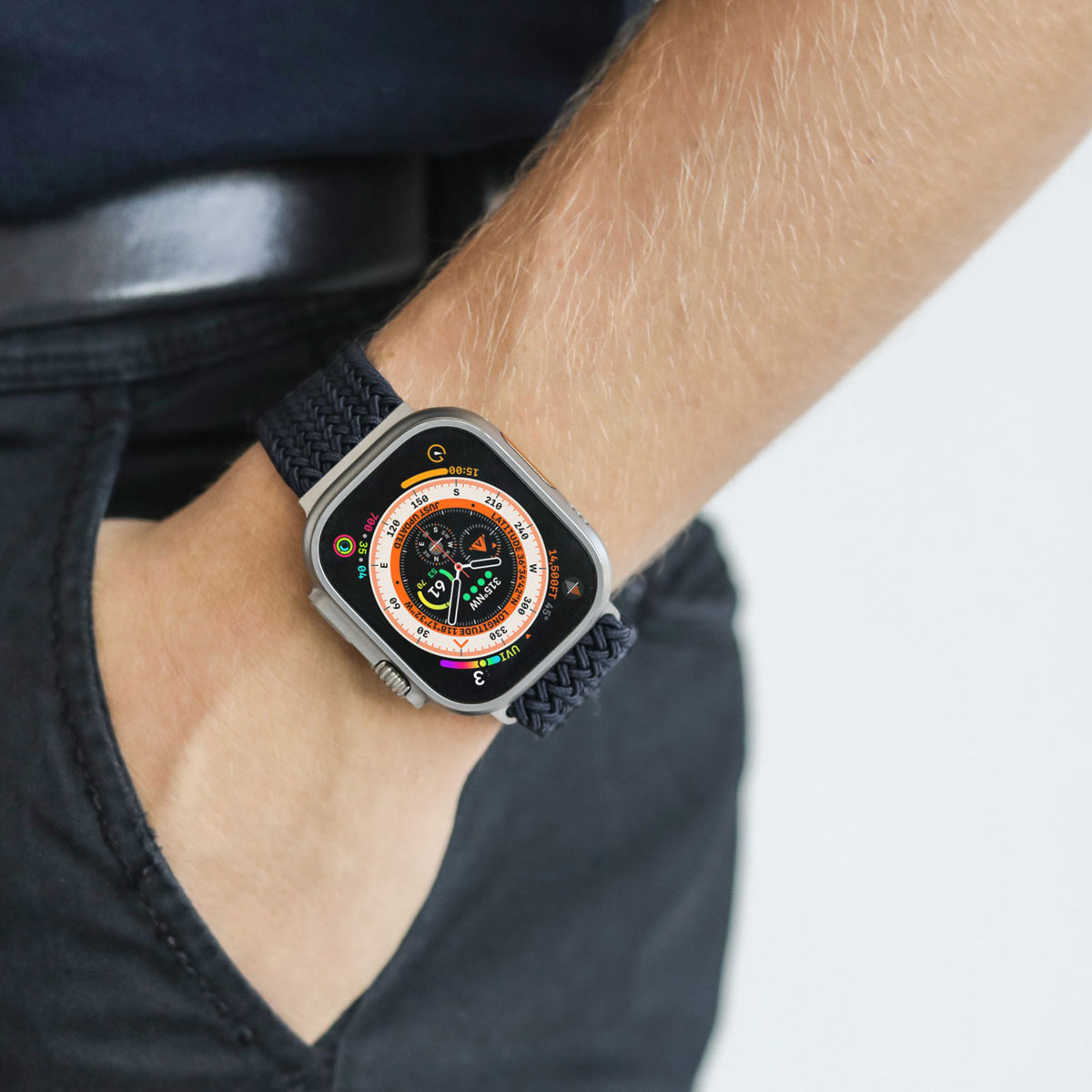 Apple Watch Ultra z czarnym plecionym paskiem na nadgarstku osoby w ciemnych spodniach.