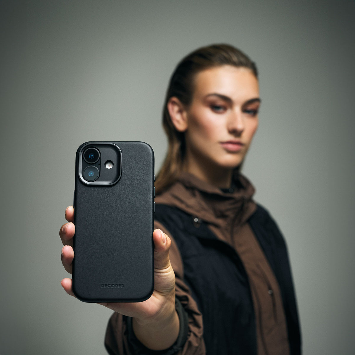 Una mujer sostiene un teléfono inteligente negro con funda de cuero frente a un fondo gris.